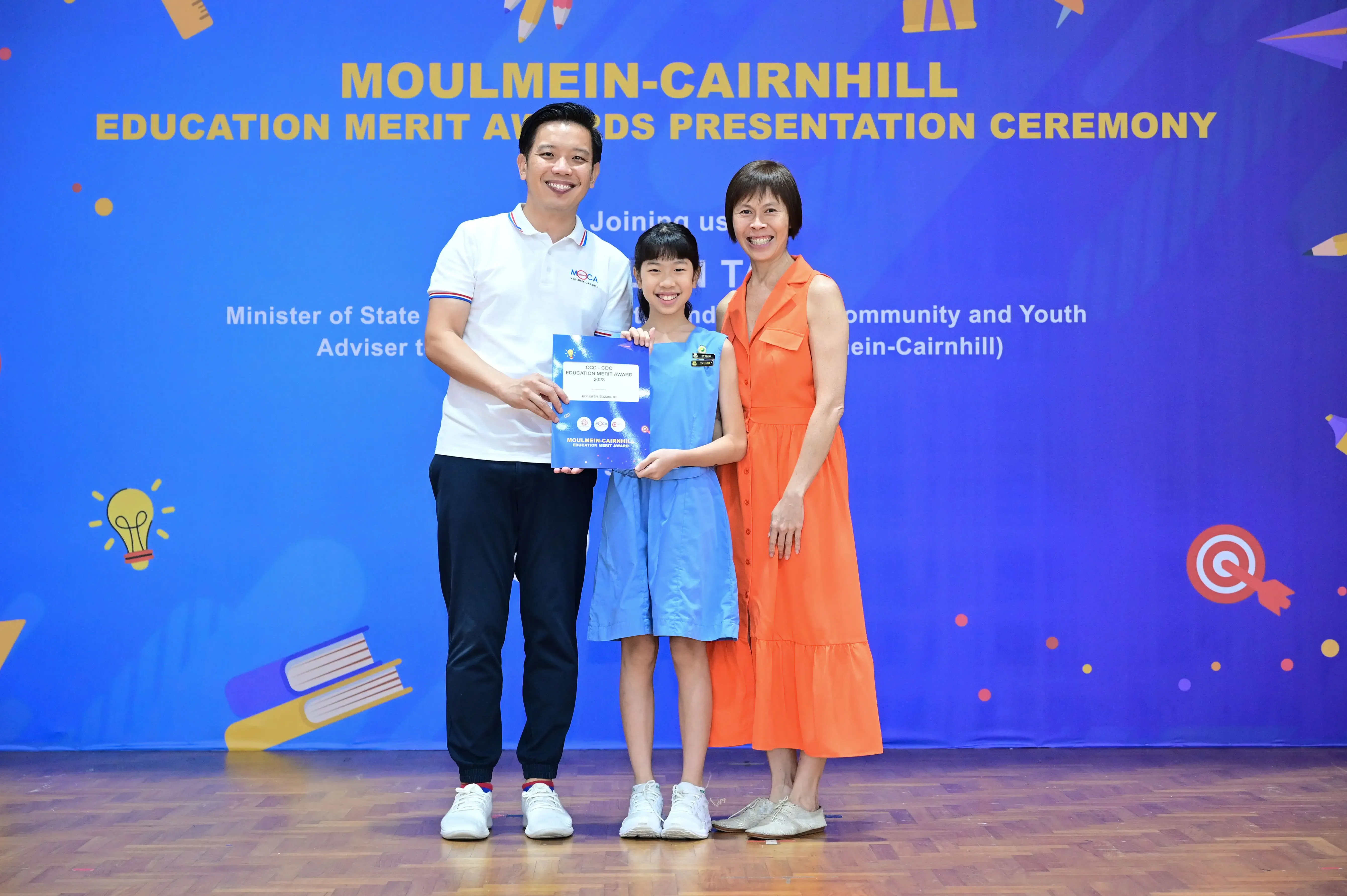 Three people pose at the Moulmein-Cairnhill Education Merit Awards Presentation Ceremony, a girl holding a blue award certificate.