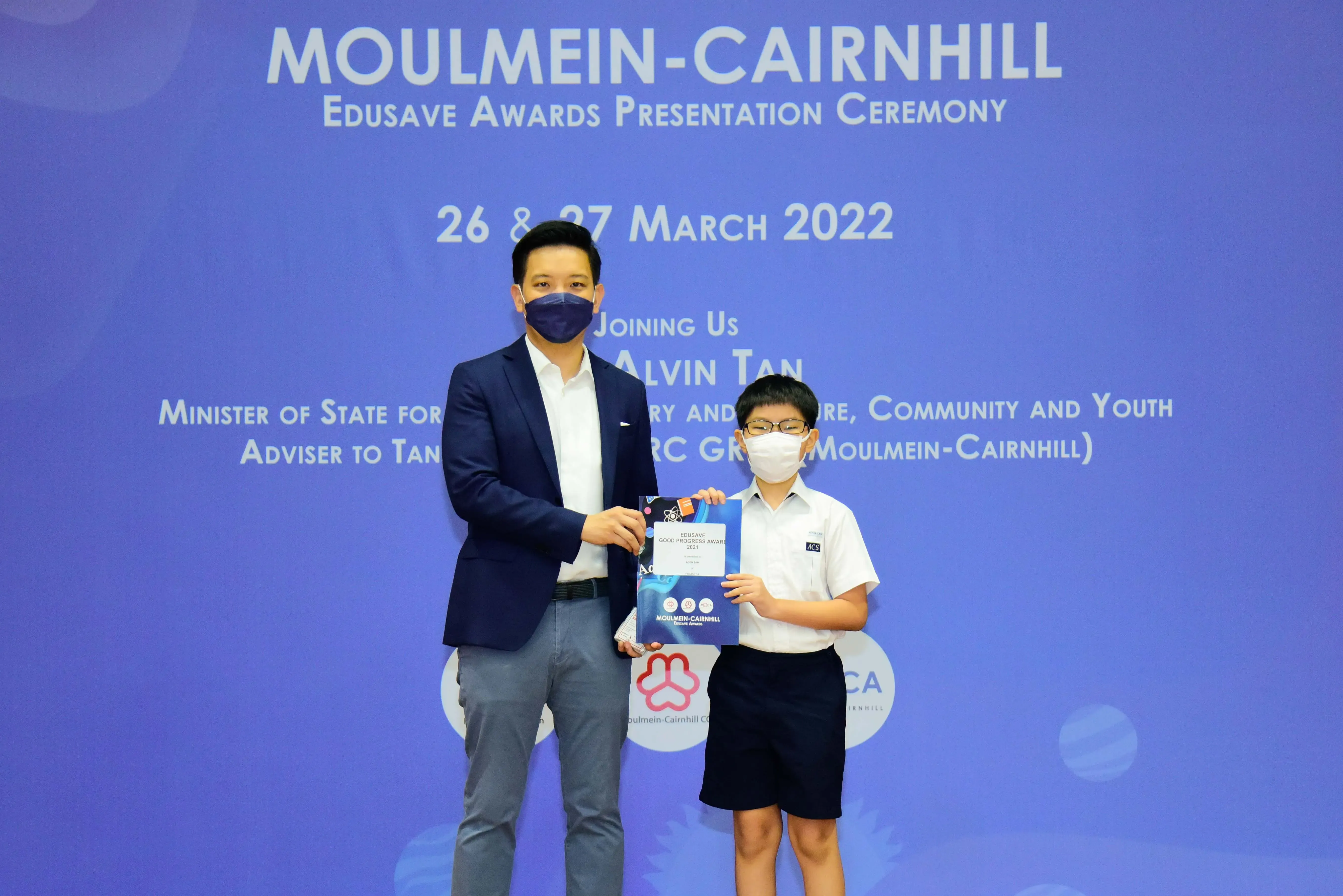 An adult in a suit and a young student in uniform hold an award at the Moulmein-Cairnhill Edusave ceremony.