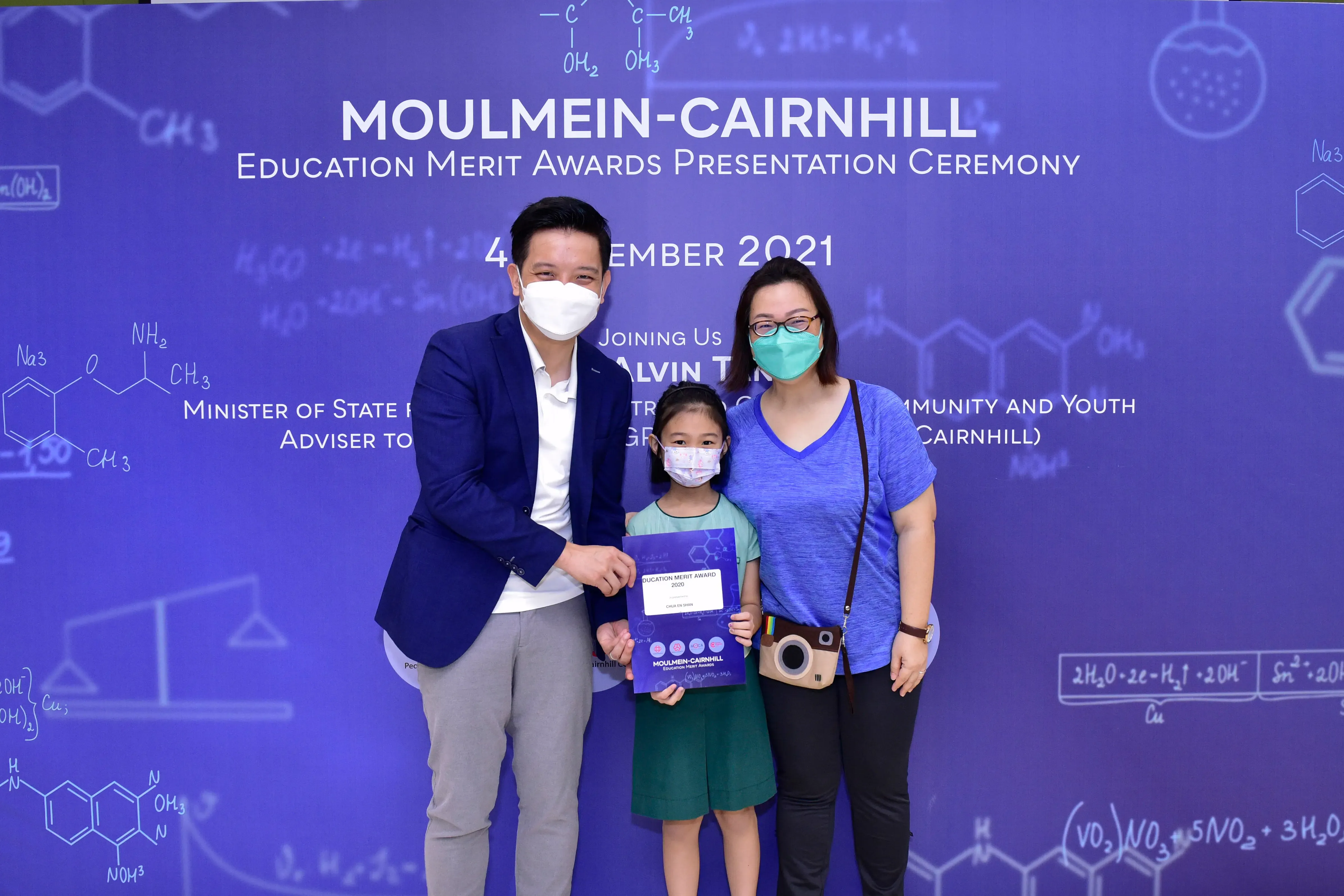 Three masked people pose at the Moulmein-Cairnhill Education Merit Awards 2021, with a child holding a certificate.