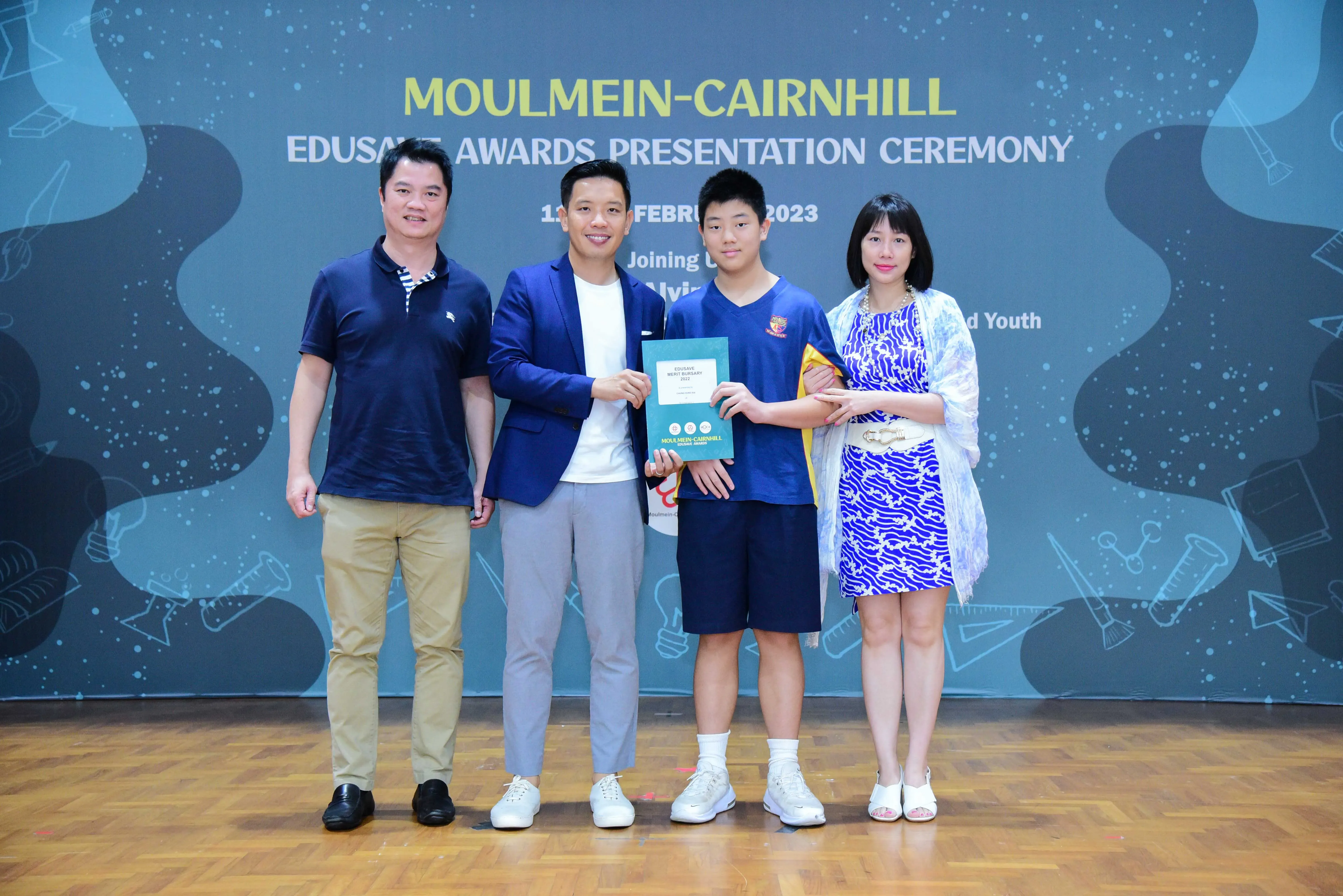 Four people stand on a wooden stage holding a certificate at the Moulmein-Cairnhill Edusave Awards Ceremony 2023.