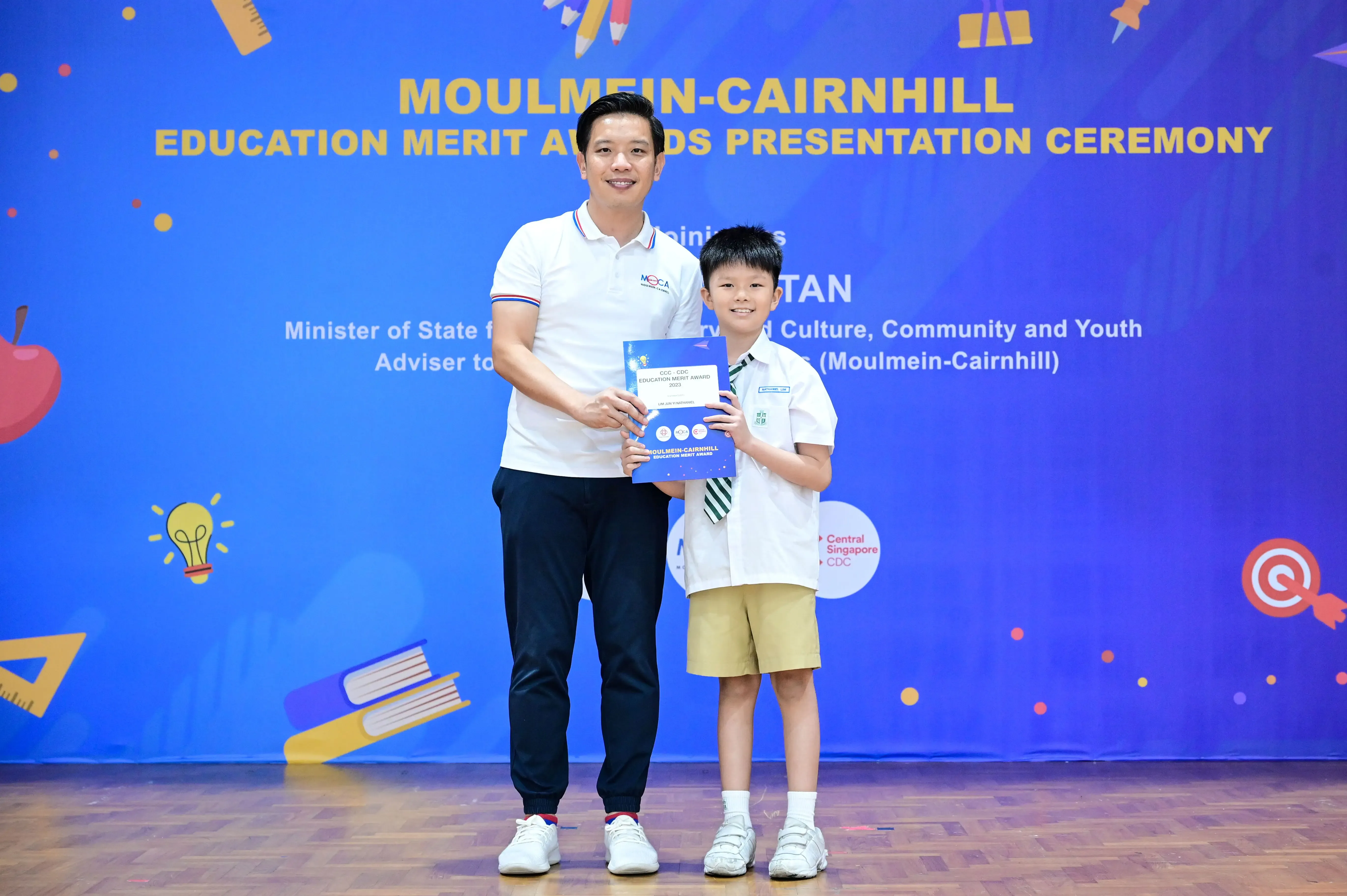 An adult and a young boy hold an award certificate on stage at the Moulmein-Cairnhill Education Merit Awards ceremony.