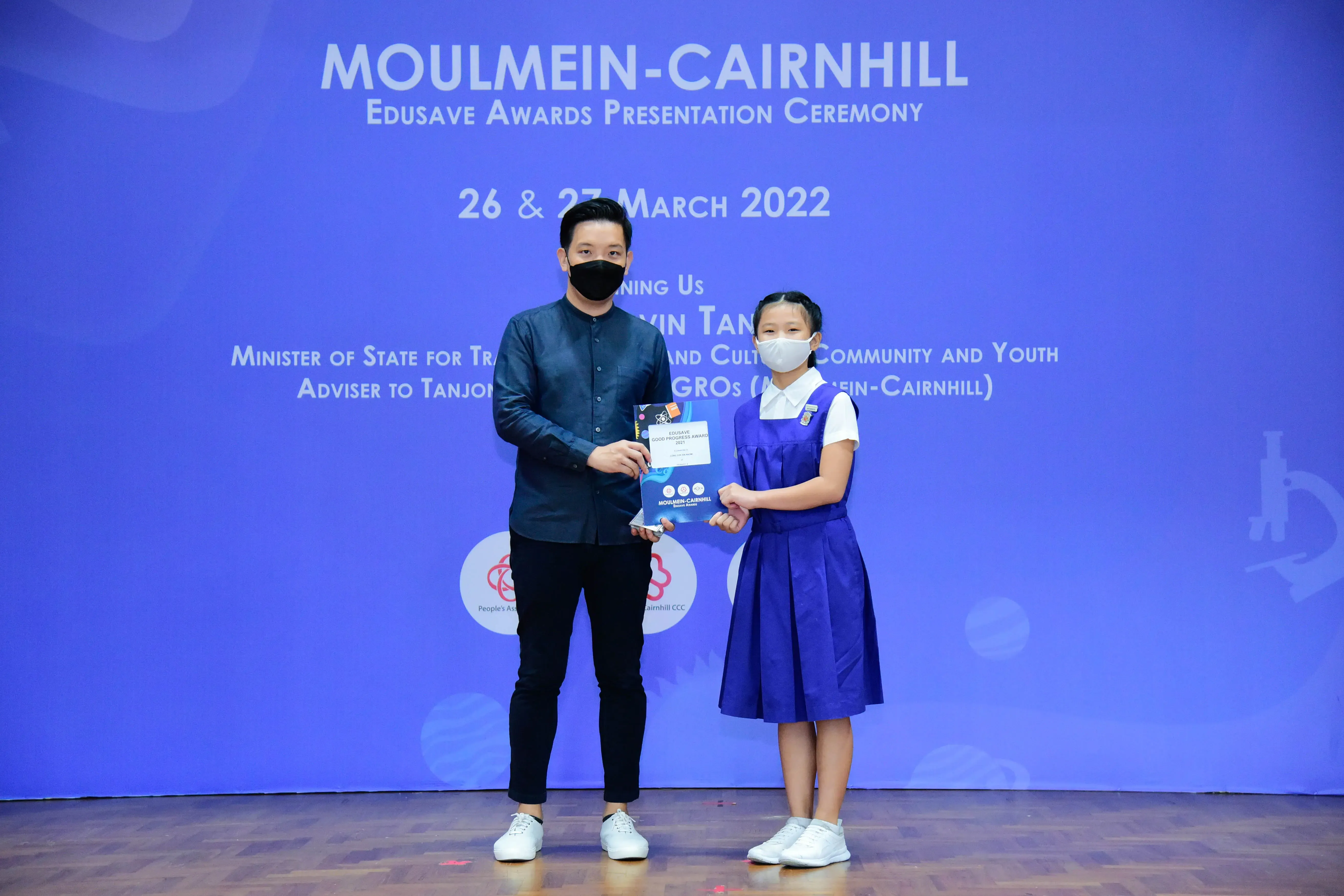 A masked man and a young girl in school uniform hold an award at the Moulmein-Cairnhill Edusave Awards Ceremony 2022.