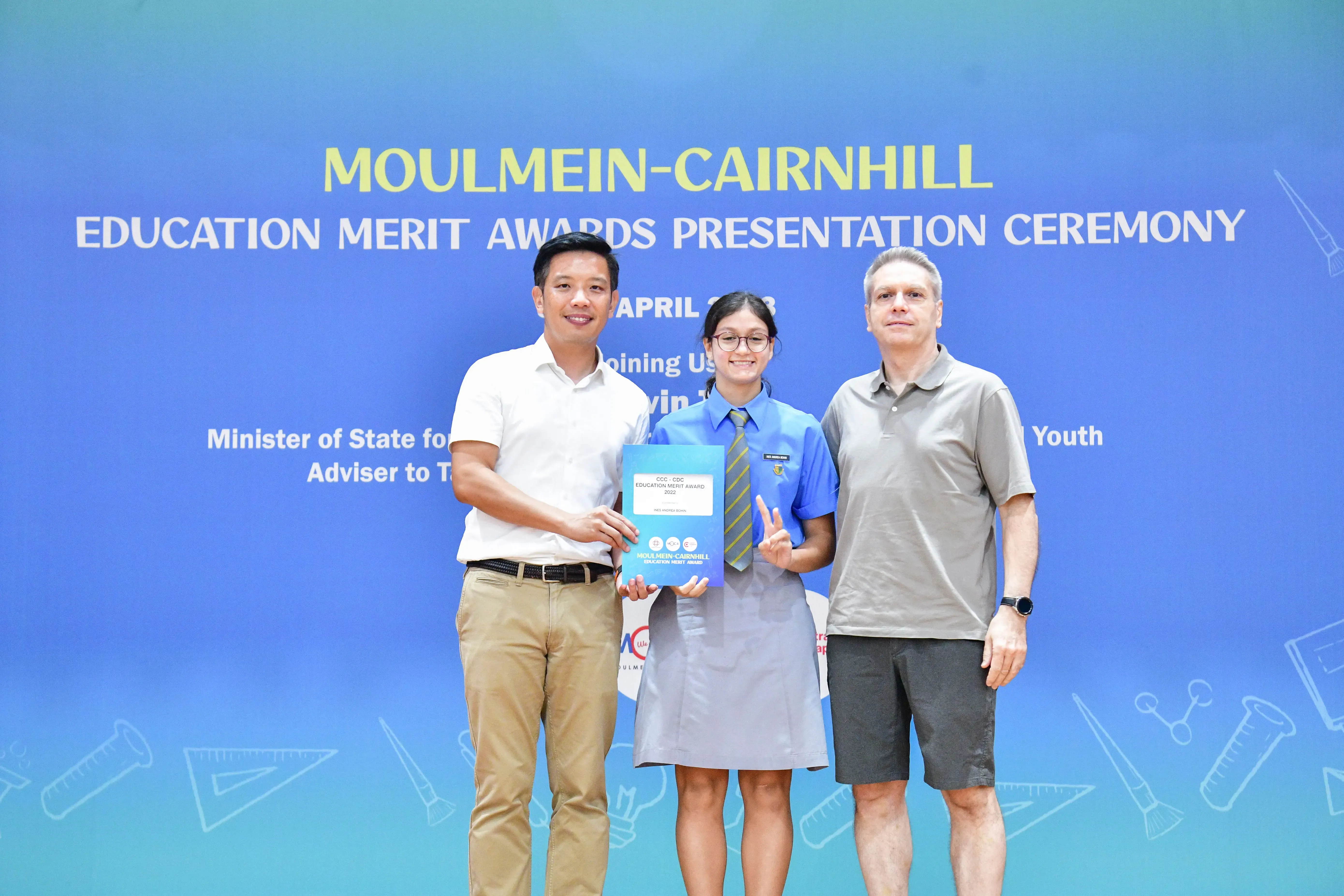A student in school uniform holds an award certificate flanked by two adults at a Moulmein-Cairnhill Education Merit Awards ceremony.