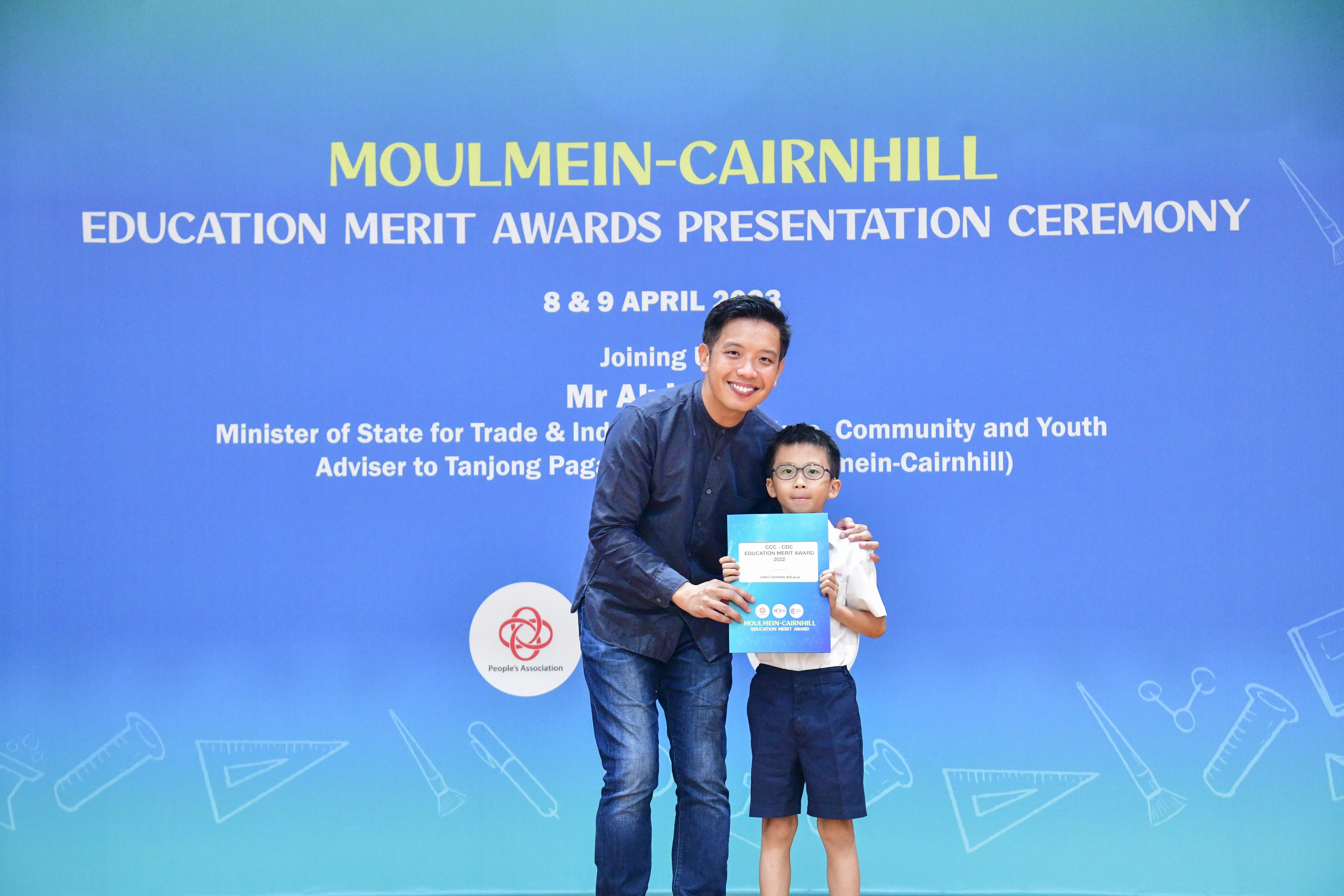 Adult man and young boy holding an award certificate at the Moulmein-Cairnhill Education Merit Awards ceremony.