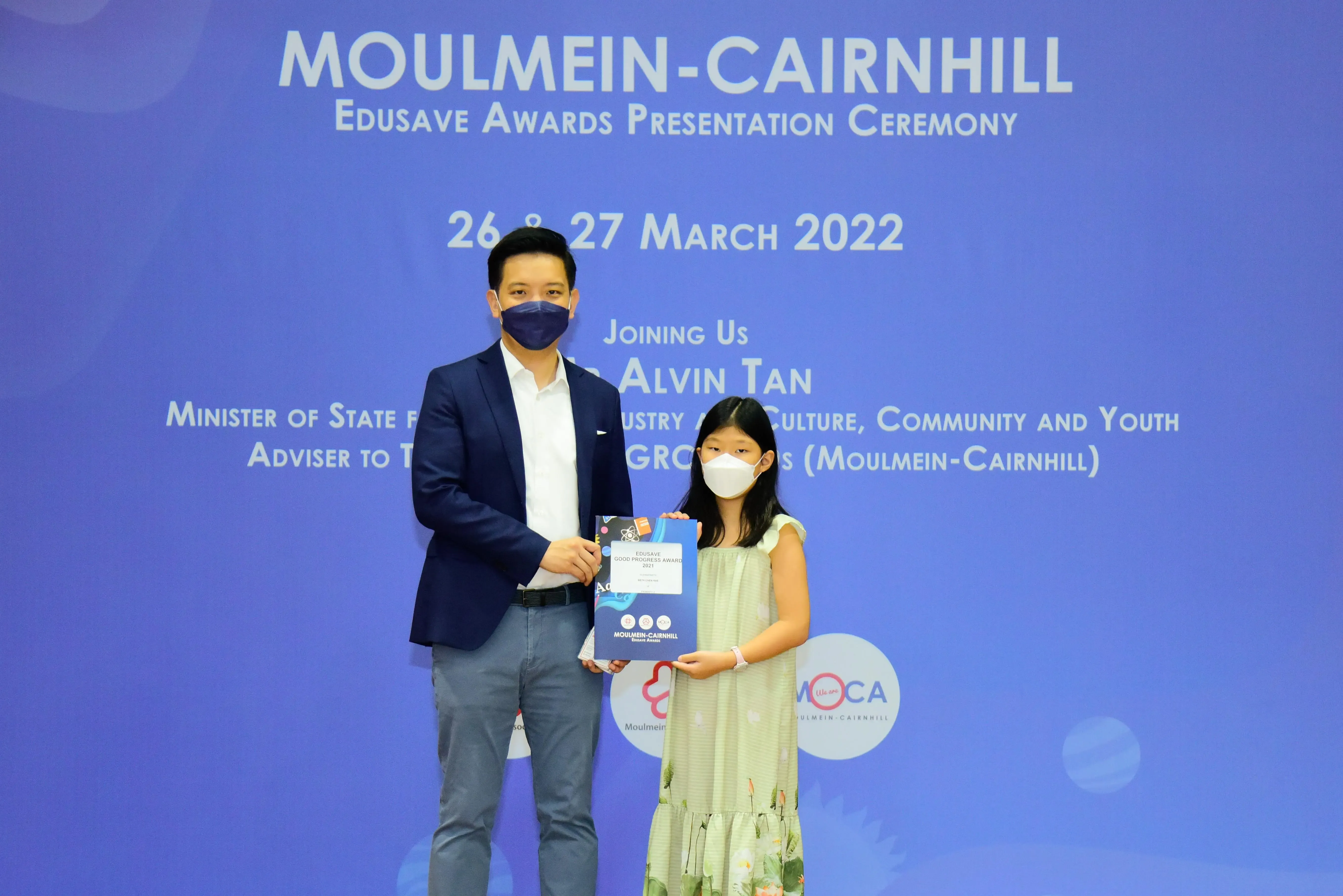 A masked man in a suit and a young girl in a dress hold an award at the Moulmein-Cairnhill Edusave Awards Ceremony.