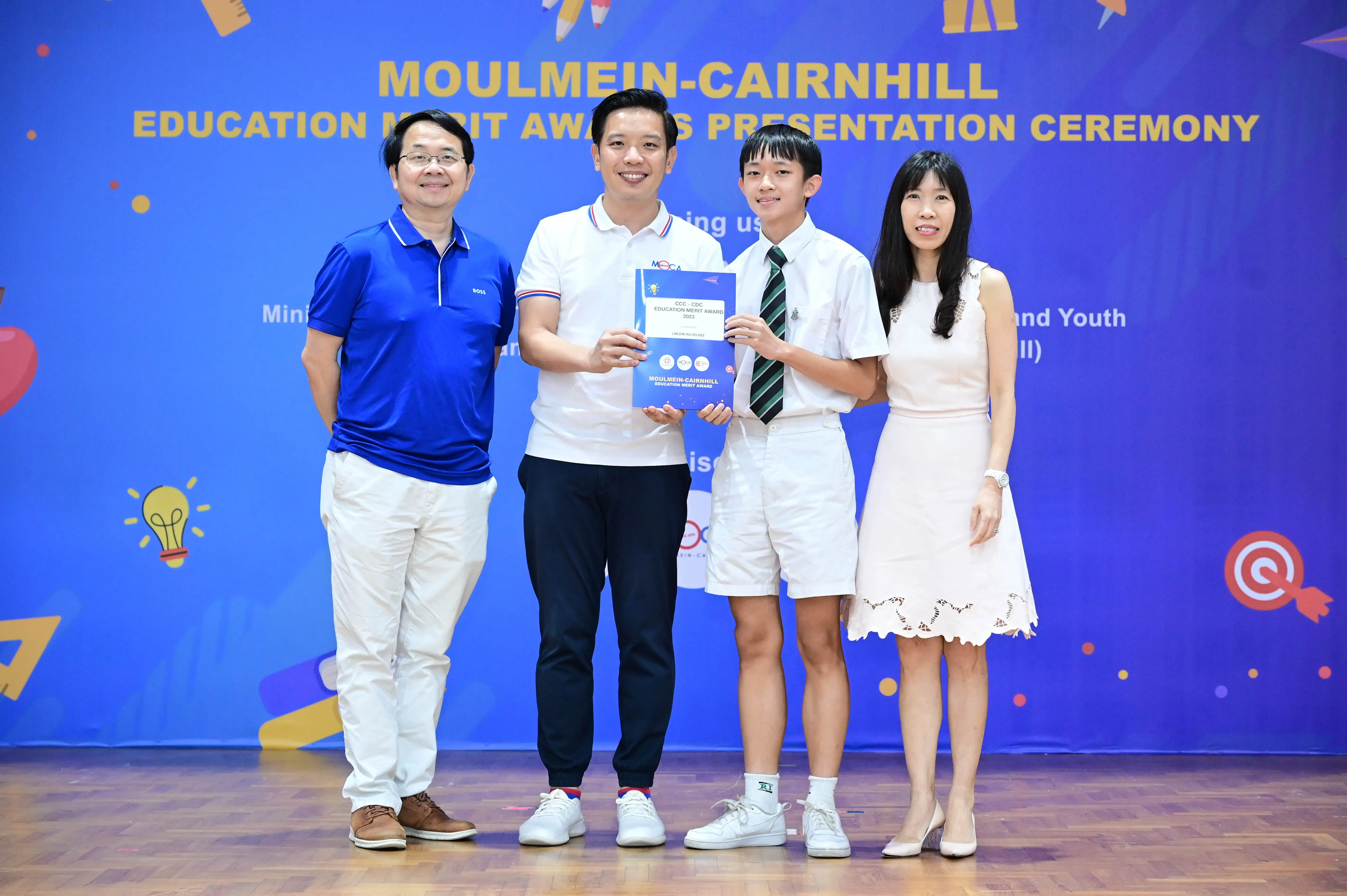 Four people pose at the Moulmein-Cairnhill Education Merit Awards Presentation Ceremony, with a student holding a certificate.