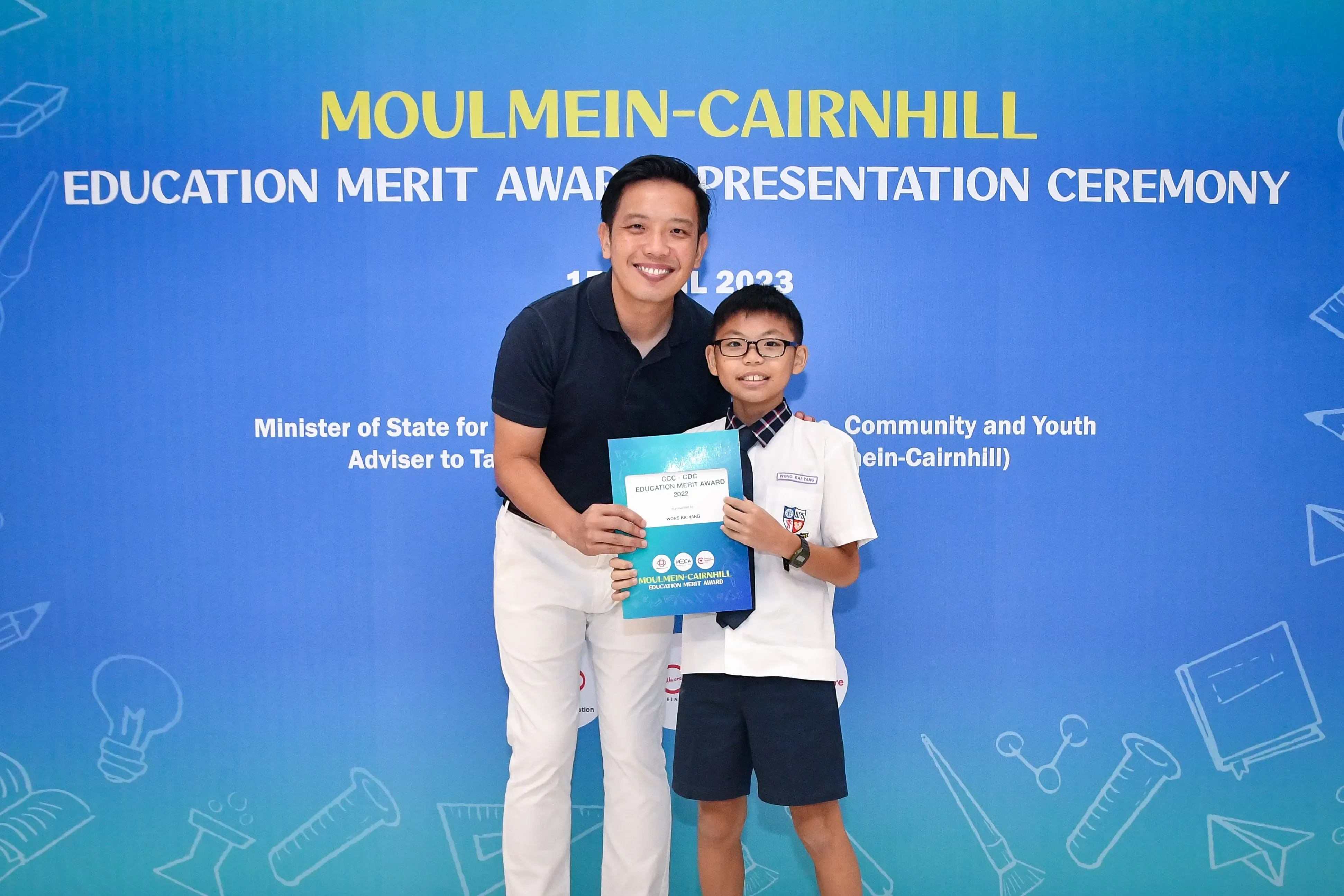 A man and a young boy in school uniform hold an award folder at the Moulmein-Cairnhill Education Merit Award Ceremony 2023.