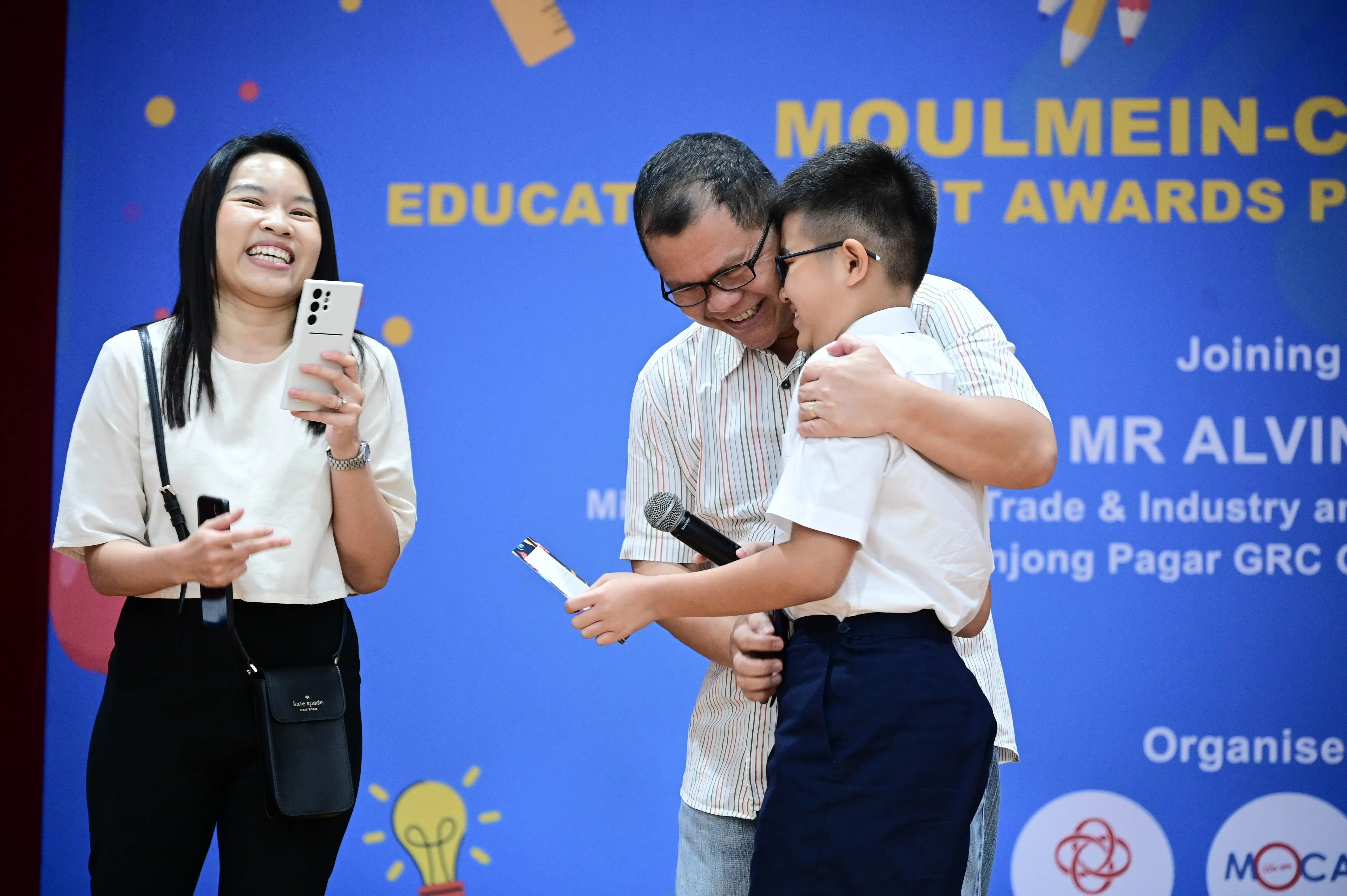 A man hugs a young boy on stage at an education awards event while a woman smiles and takes a photo nearby.