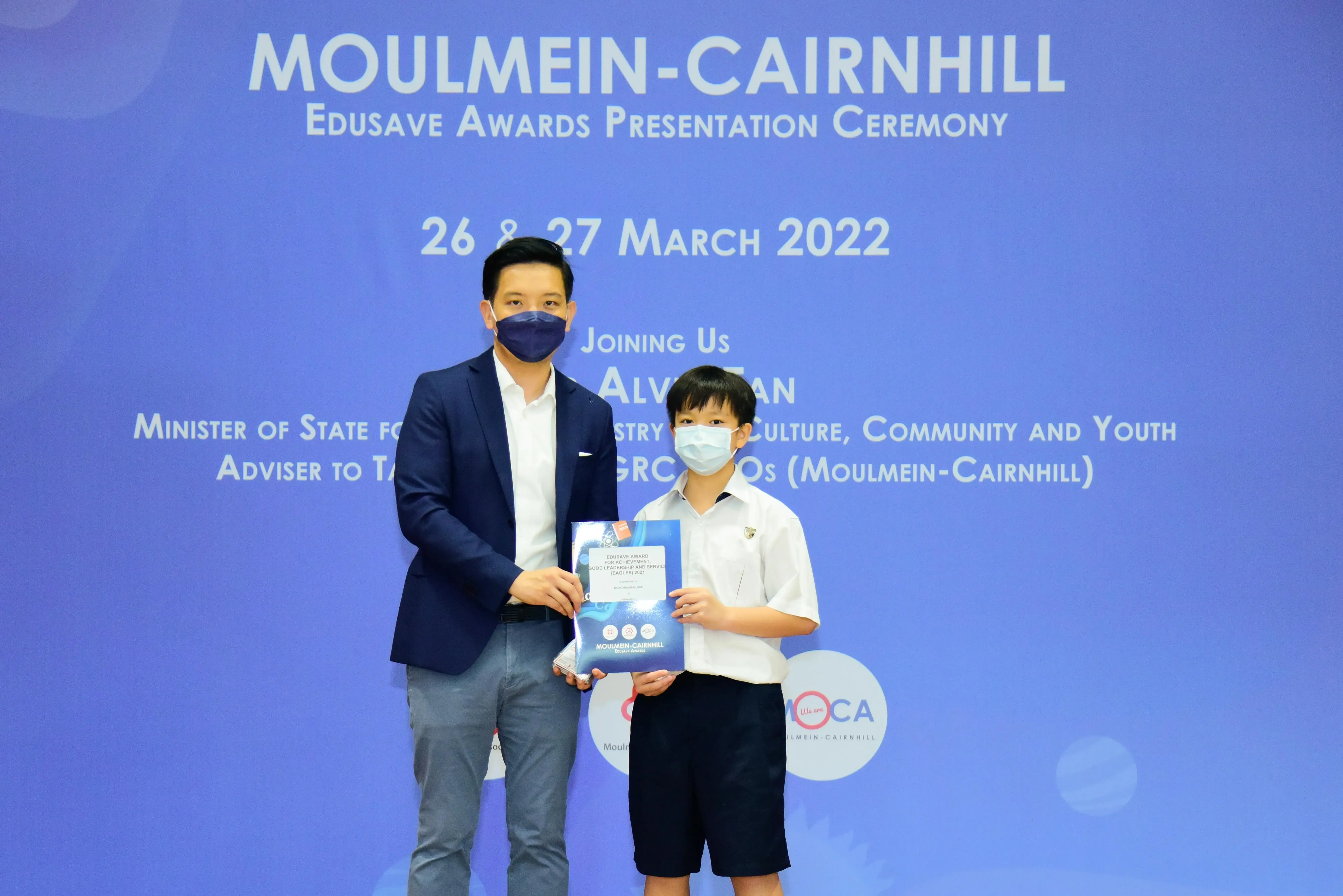 An adult and a young student in school uniform hold an award together at the Moulmein-Cairnhill Edusave Awards Ceremony 2022.