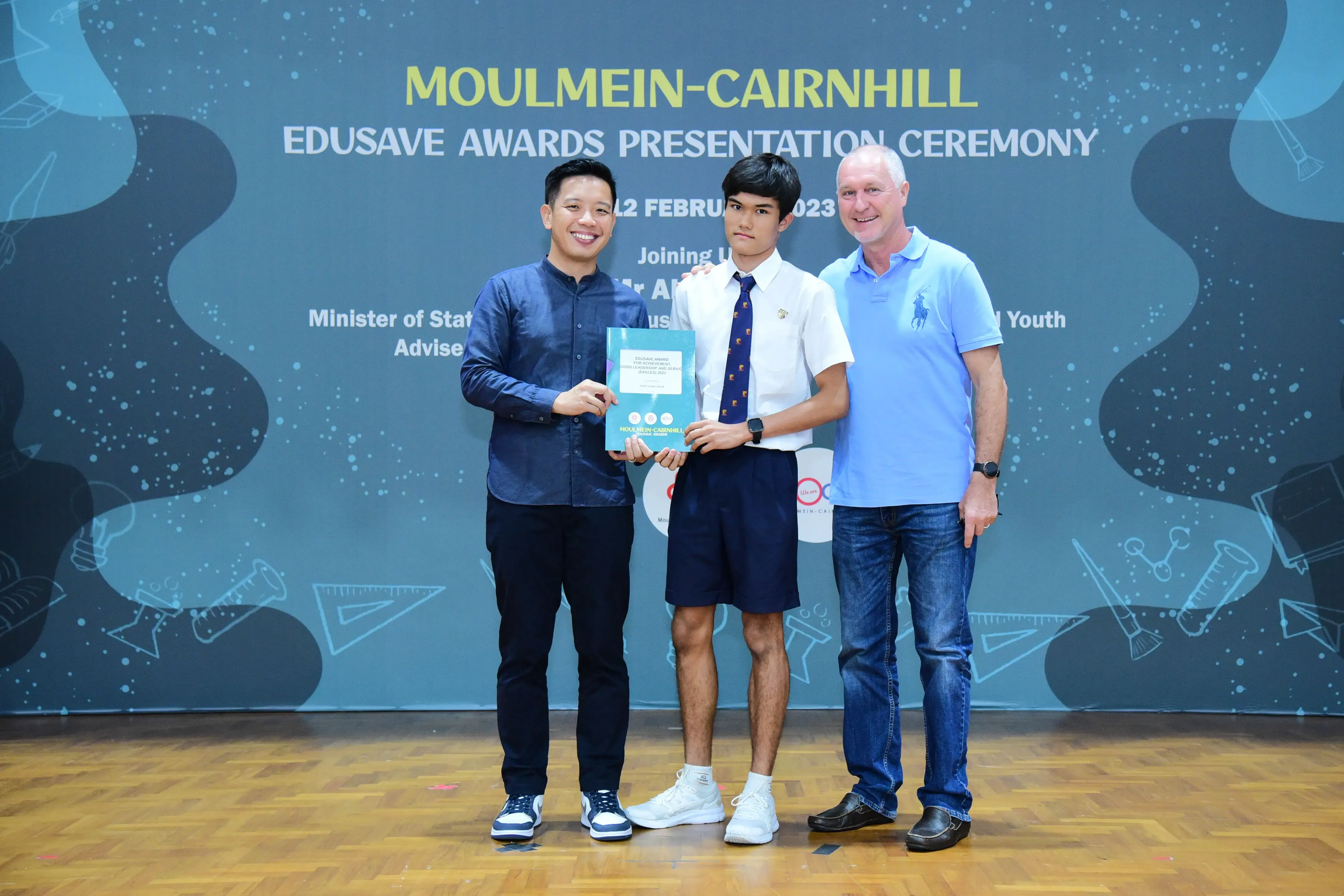 Three people pose on a stage at the Moulmein-Cairnhill Edusave Awards Presentation Ceremony, holding a certificate.