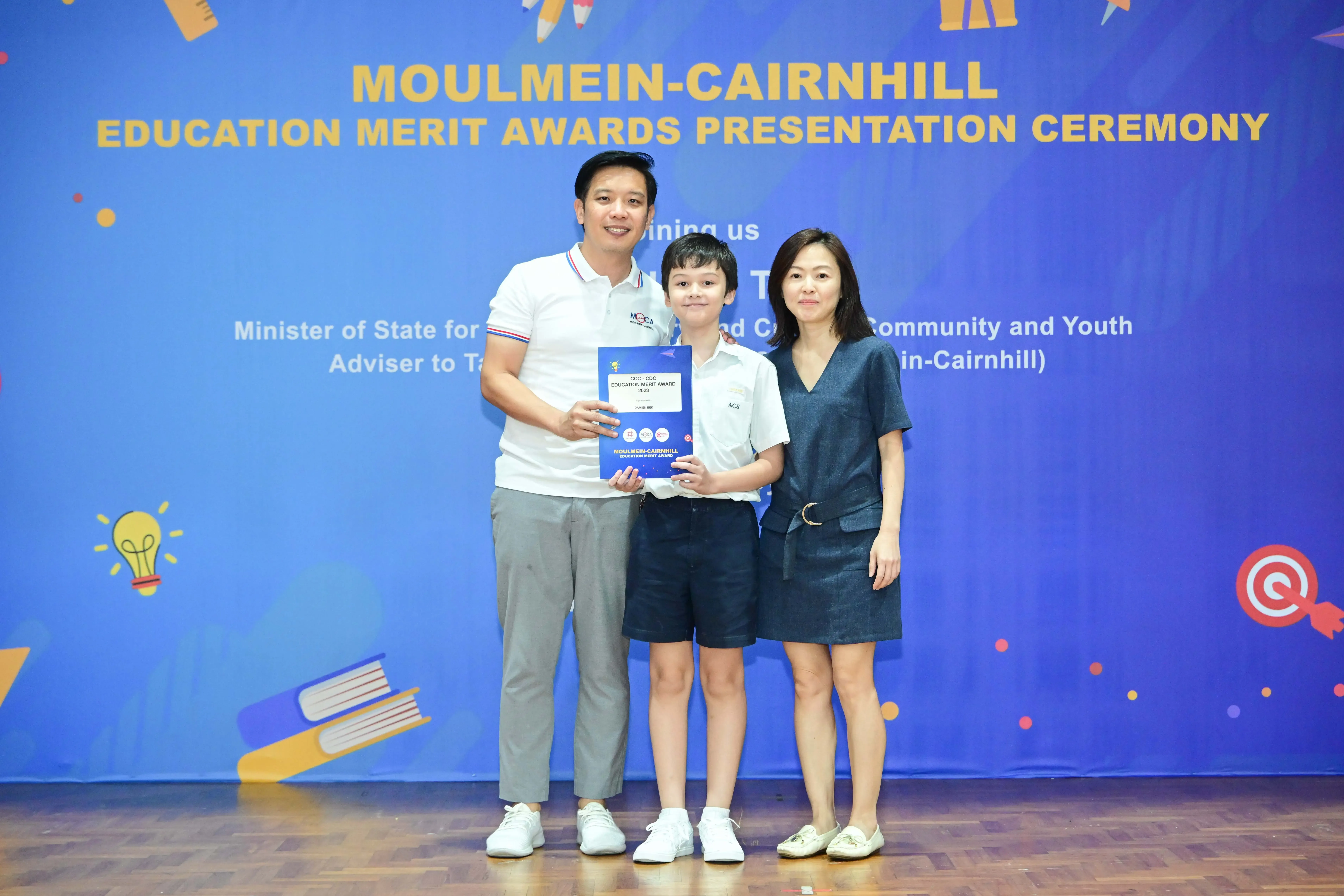 A boy holding an award certificate stands between two adults at the Moulmein-Cairnhill Education Merit Awards Ceremony.
