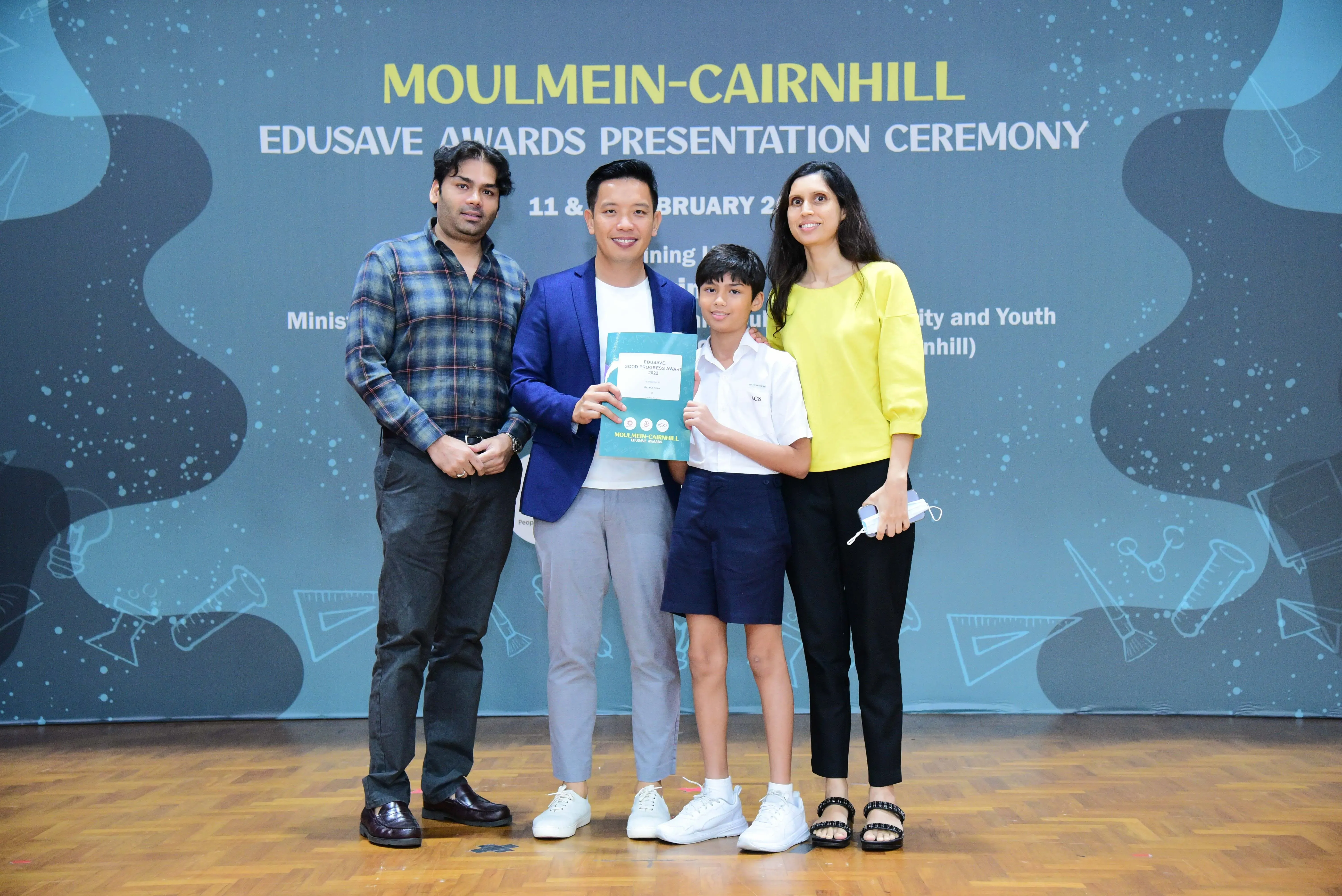 Four people pose at the Moulmein-Cairnhill Edusave Awards Presentation Ceremony, including a child holding an award folder.