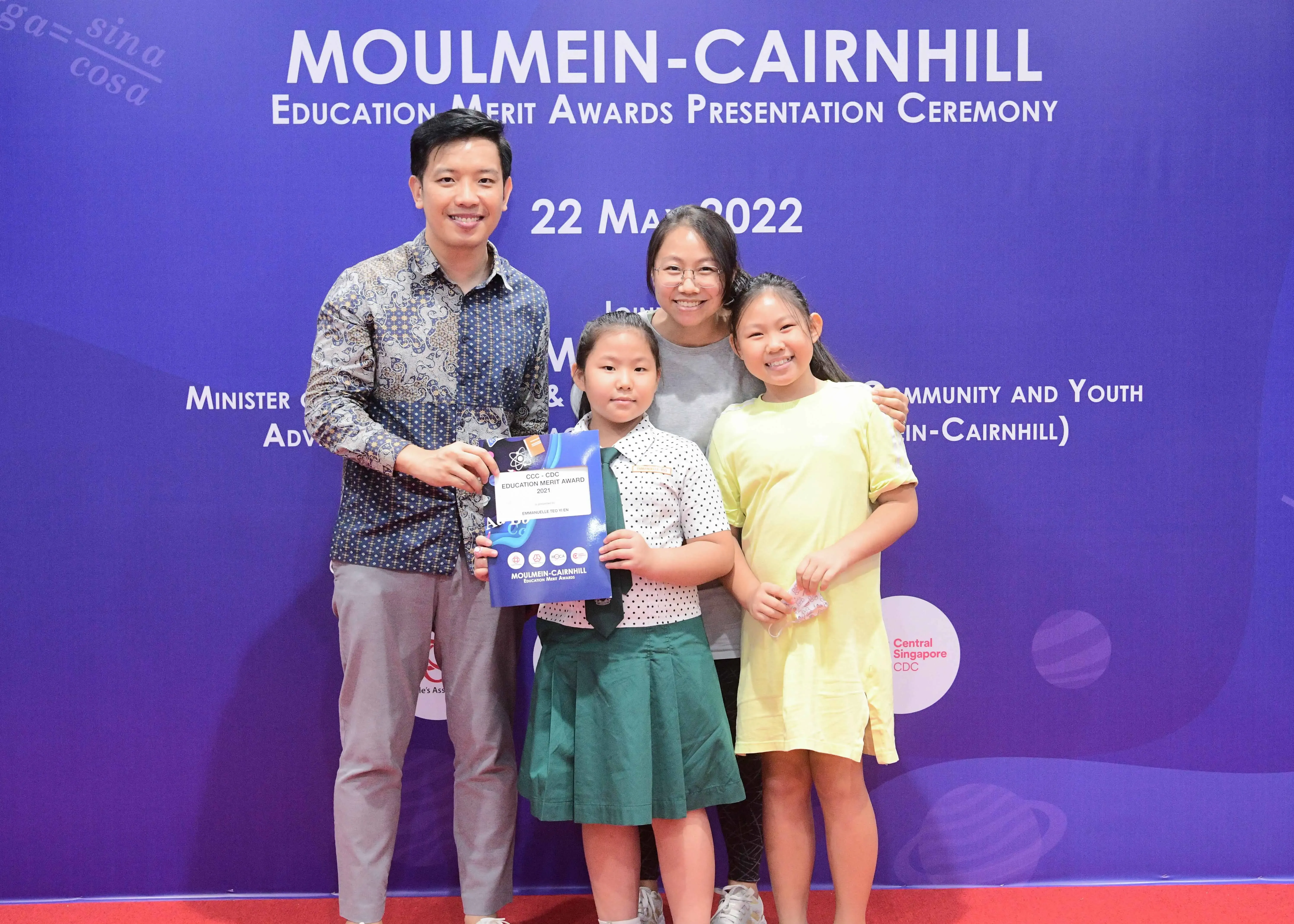 Four people pose smiling before a Moulmein-Cairnhill Education Merit Awards banner dated 22 May 2022.