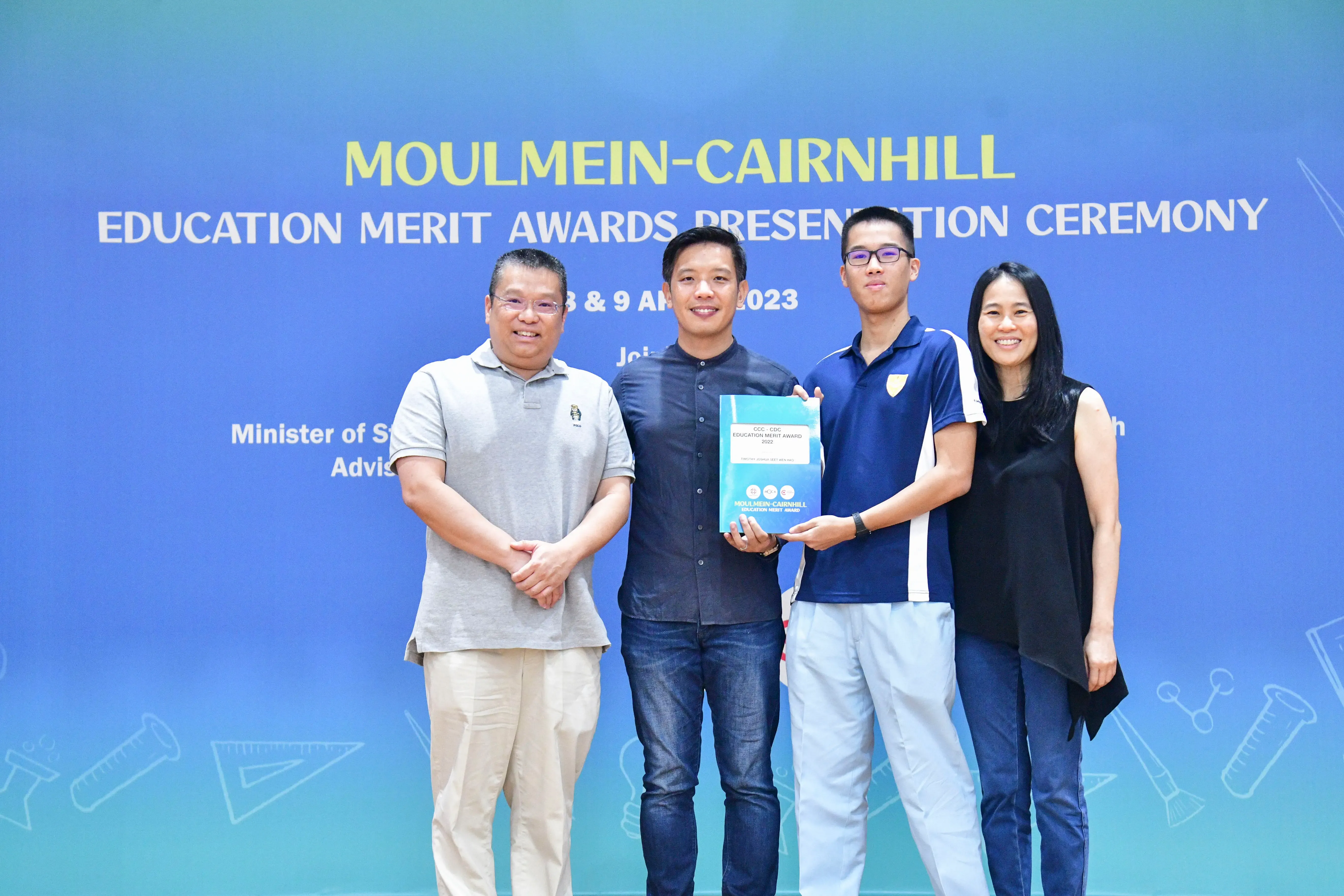 Four people pose in front of a blue Moulmein-Cairnhill Education Merit Awards Presentation Ceremony banner, holding a certificate.