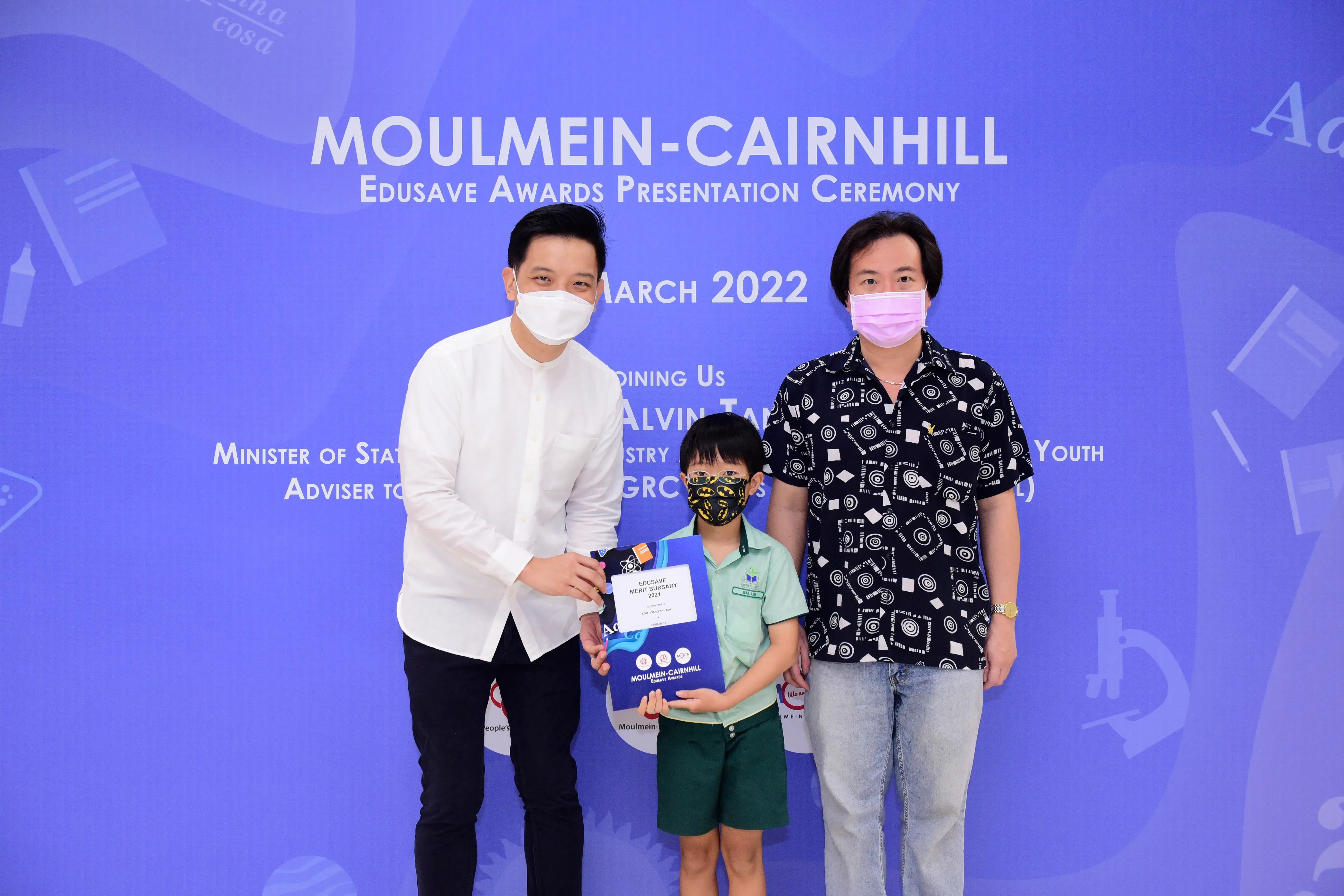 Two masked adults and a masked child holding an award at the Moulmein-Cairnhill Edusave Awards Ceremony, March 2022.