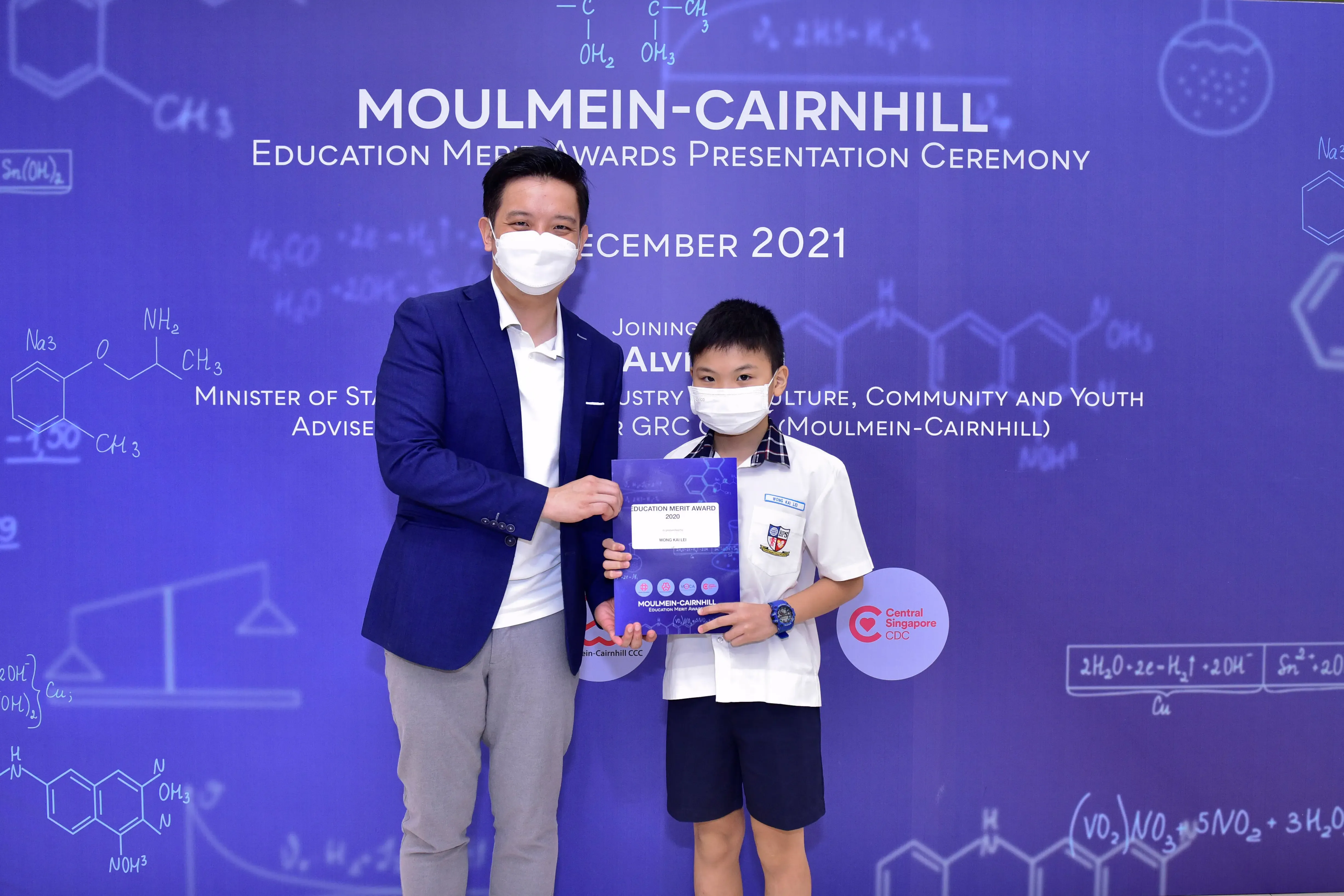 A masked man in a blazer and a young boy hold an award certificate at the Moulmein-Cairnhill Education Merit Awards 2021.
