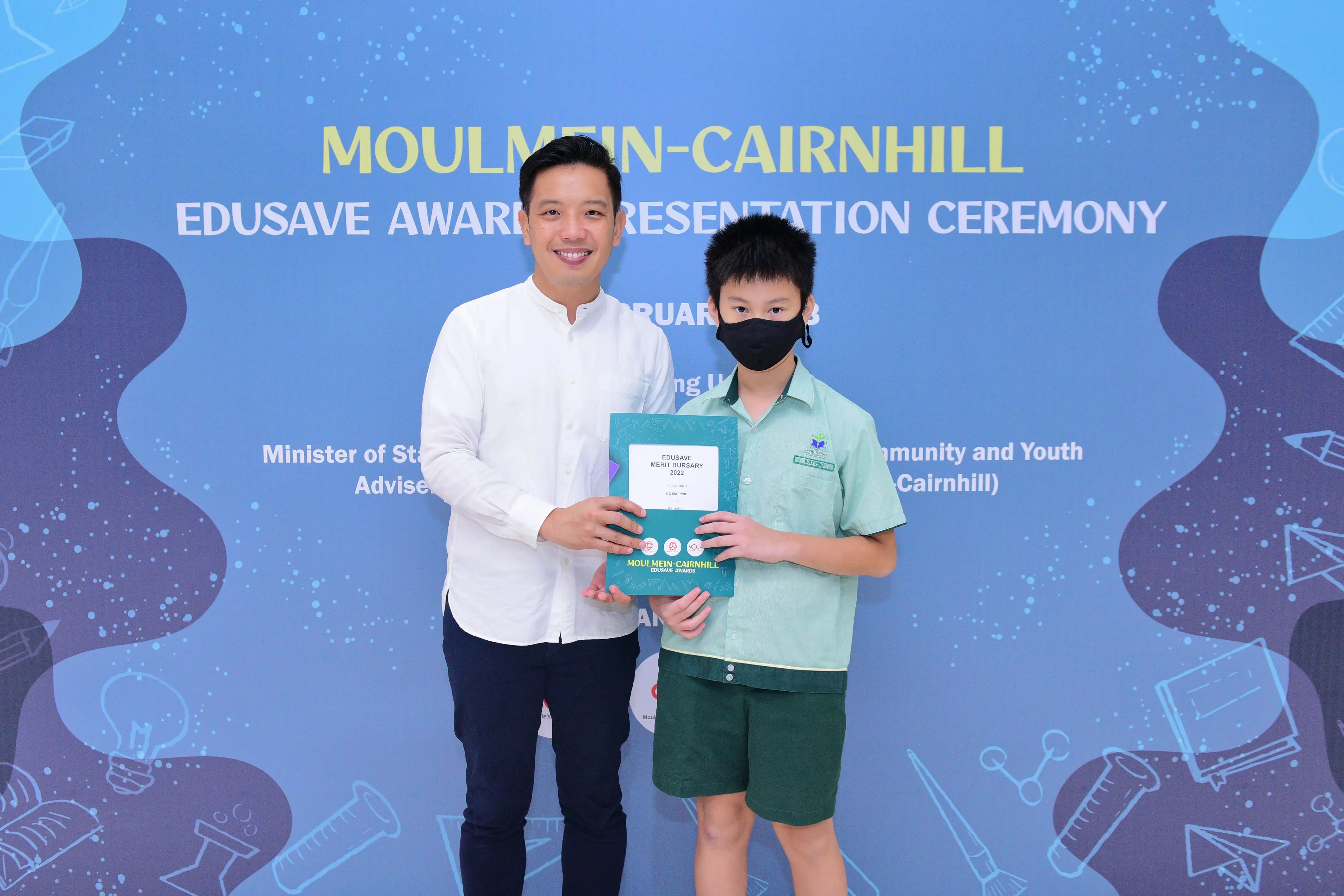 Man and masked boy hold an Edusave Merit Bursary certificate at Moulmein-Cairnhill Award Presentation Ceremony.