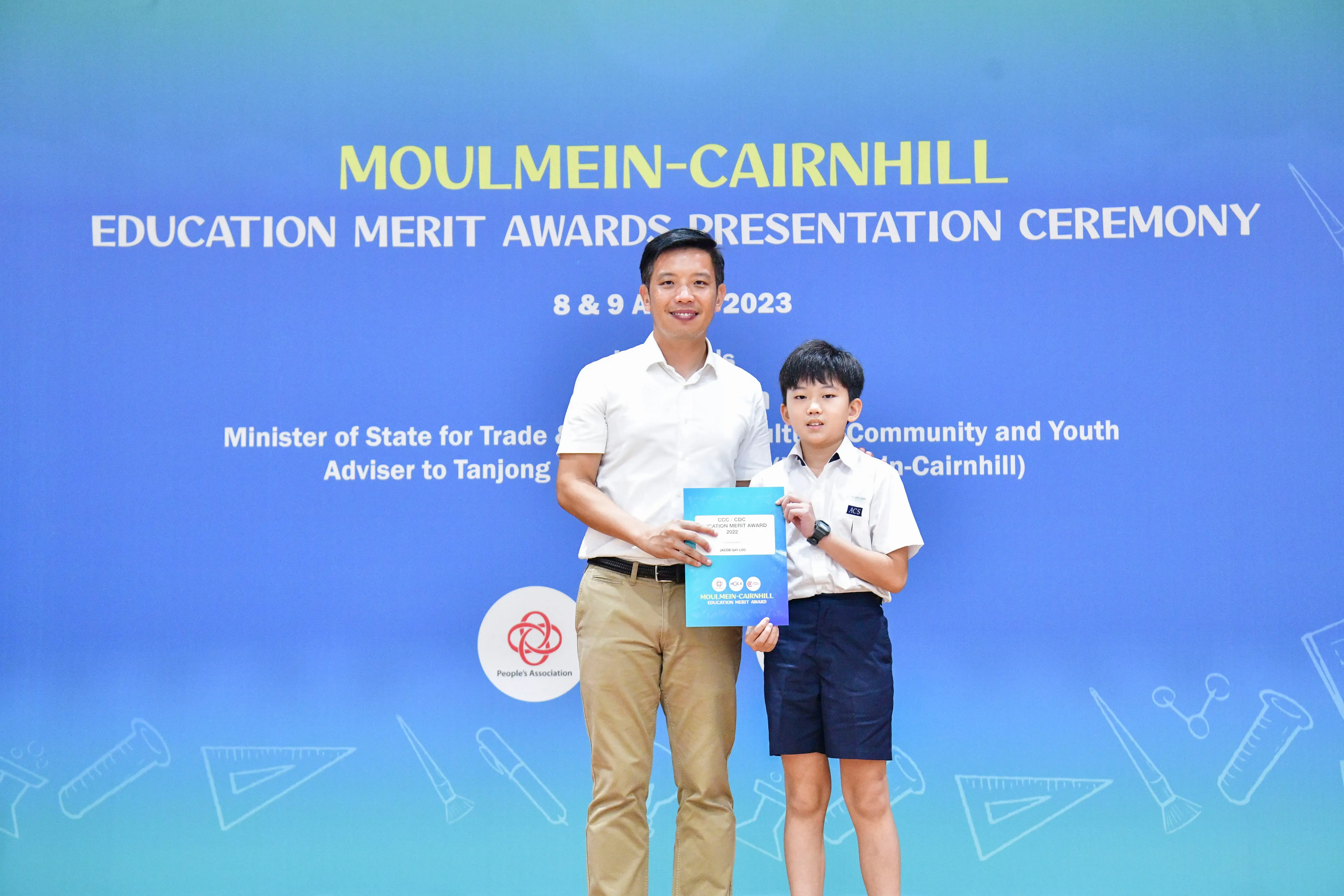 An adult man and a young boy hold award certificates in front of a Moulmein-Cairnhill Education Merit Awards banner.