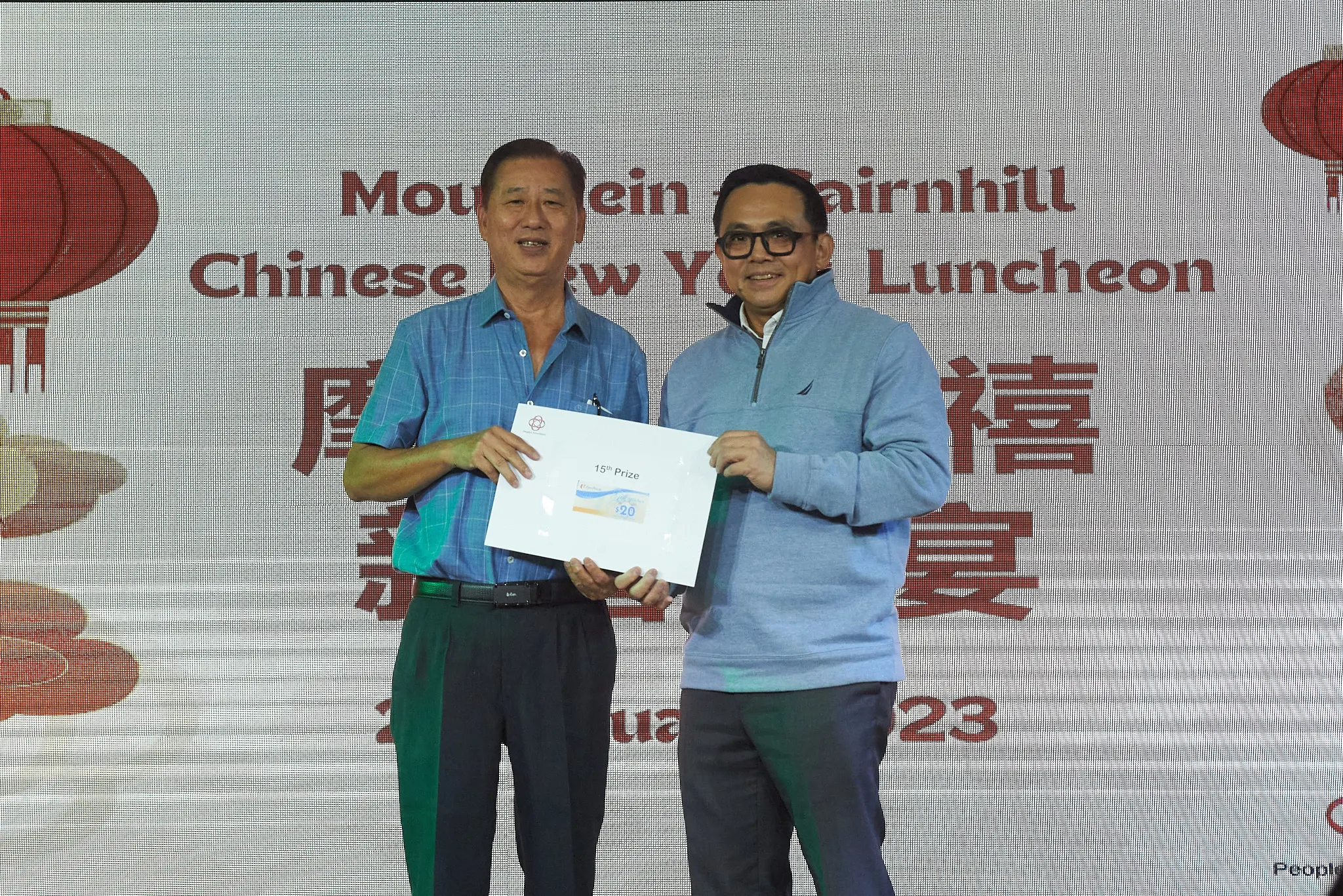 Two men hold a 15th Prize certificate at a Chinese New Year Luncheon event backdrop with red lanterns.