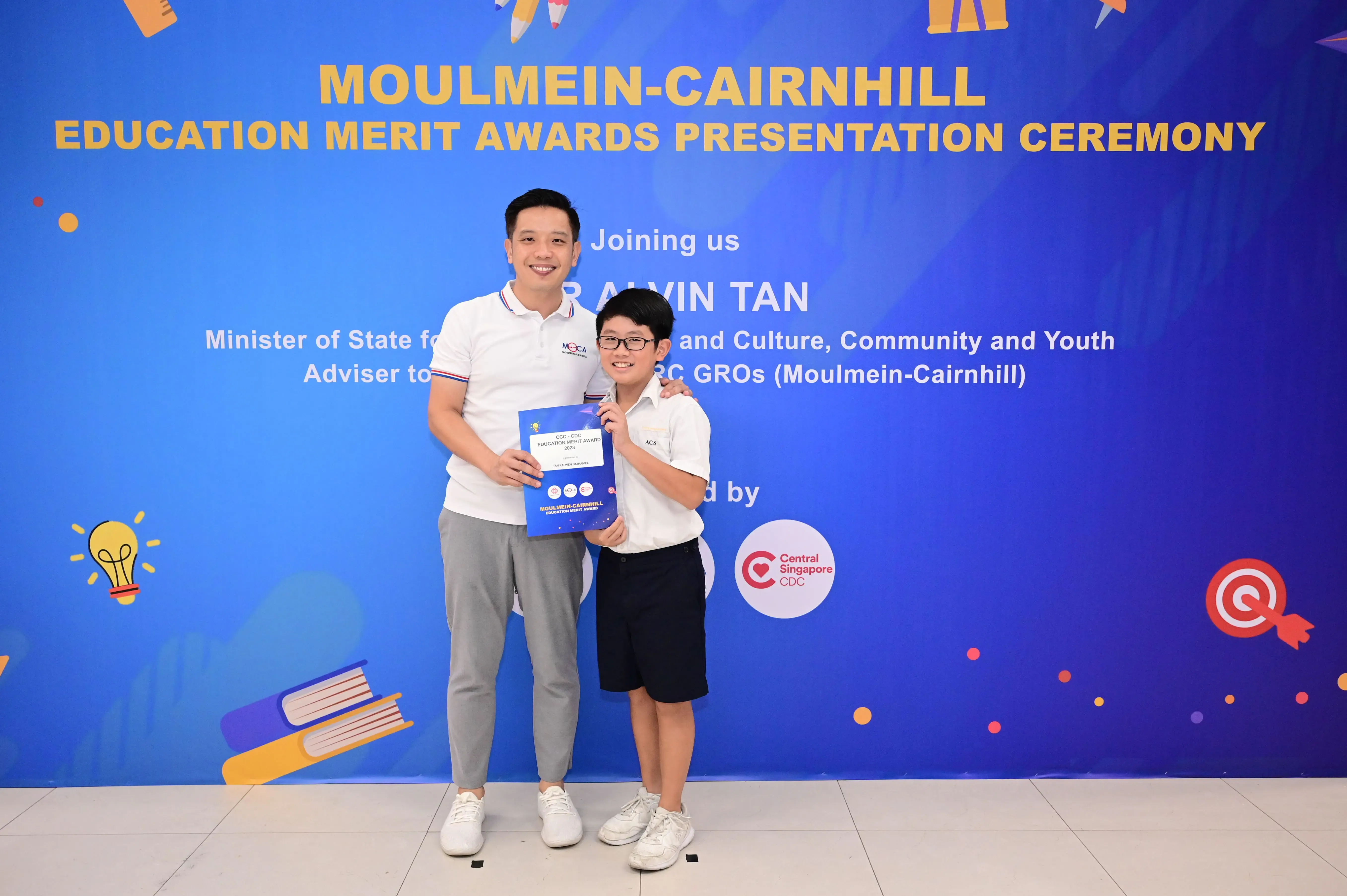 A man and a young boy holding an award certificate pose in front of a Moulmein-Cairnhill Education Merit Awards banner.
