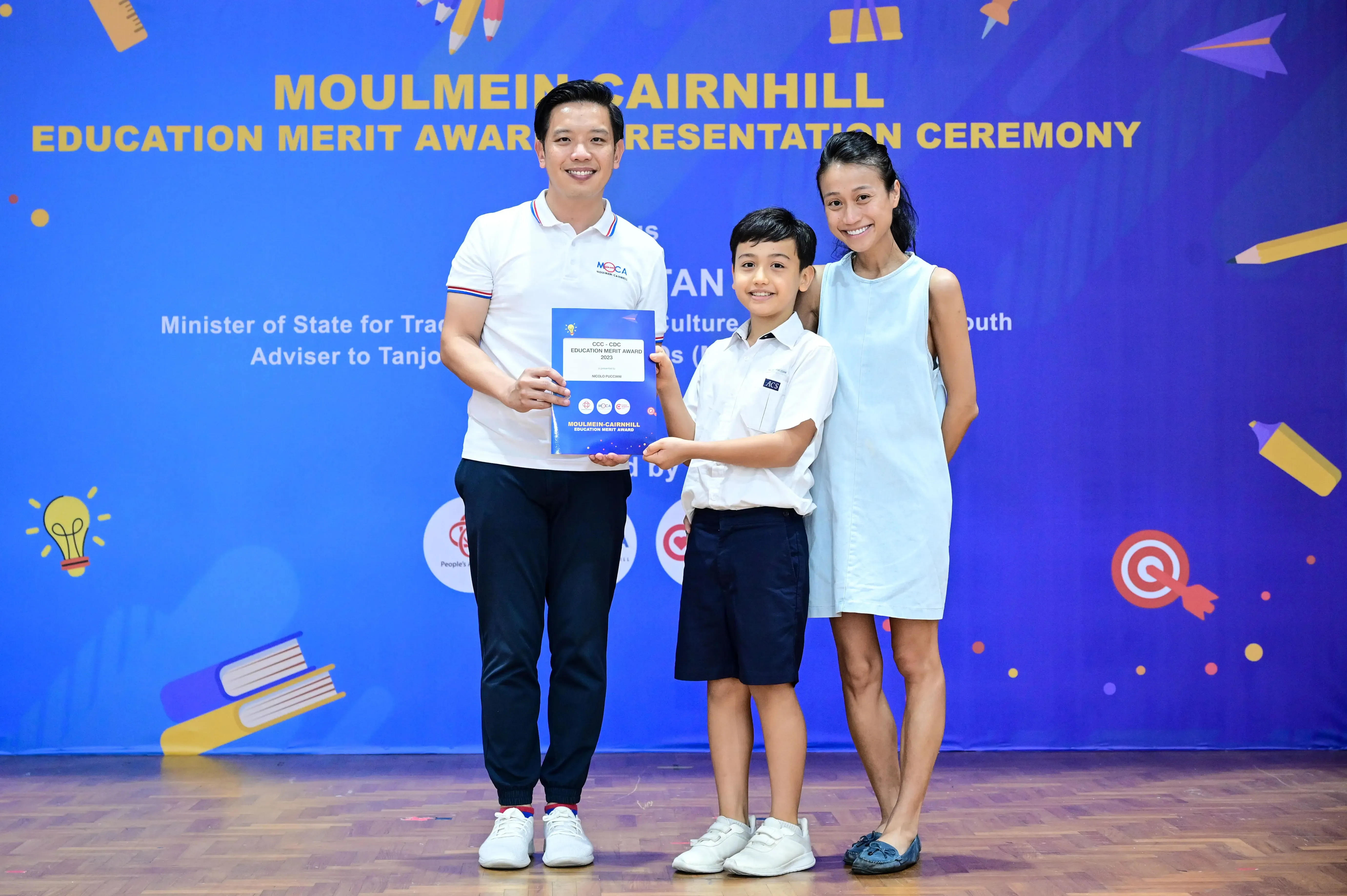 A man, boy in school uniform, and woman pose with an award at the Moulmein-Cairnhill Education Merit Award Ceremony.