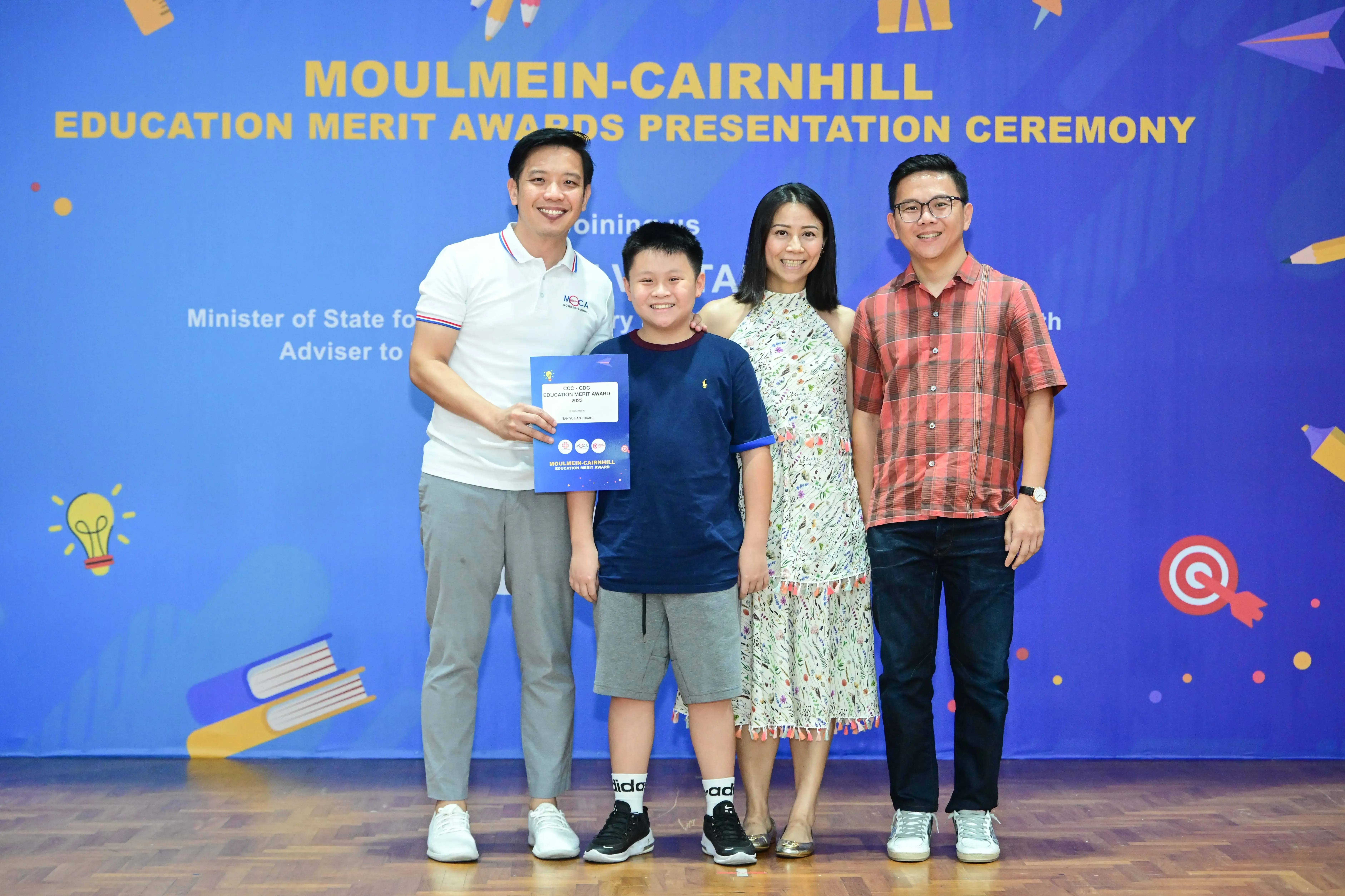 Four people pose smiling at the Moulmein-Cairnhill Education Merit Awards Presentation Ceremony, boy holds a blue award certificate.