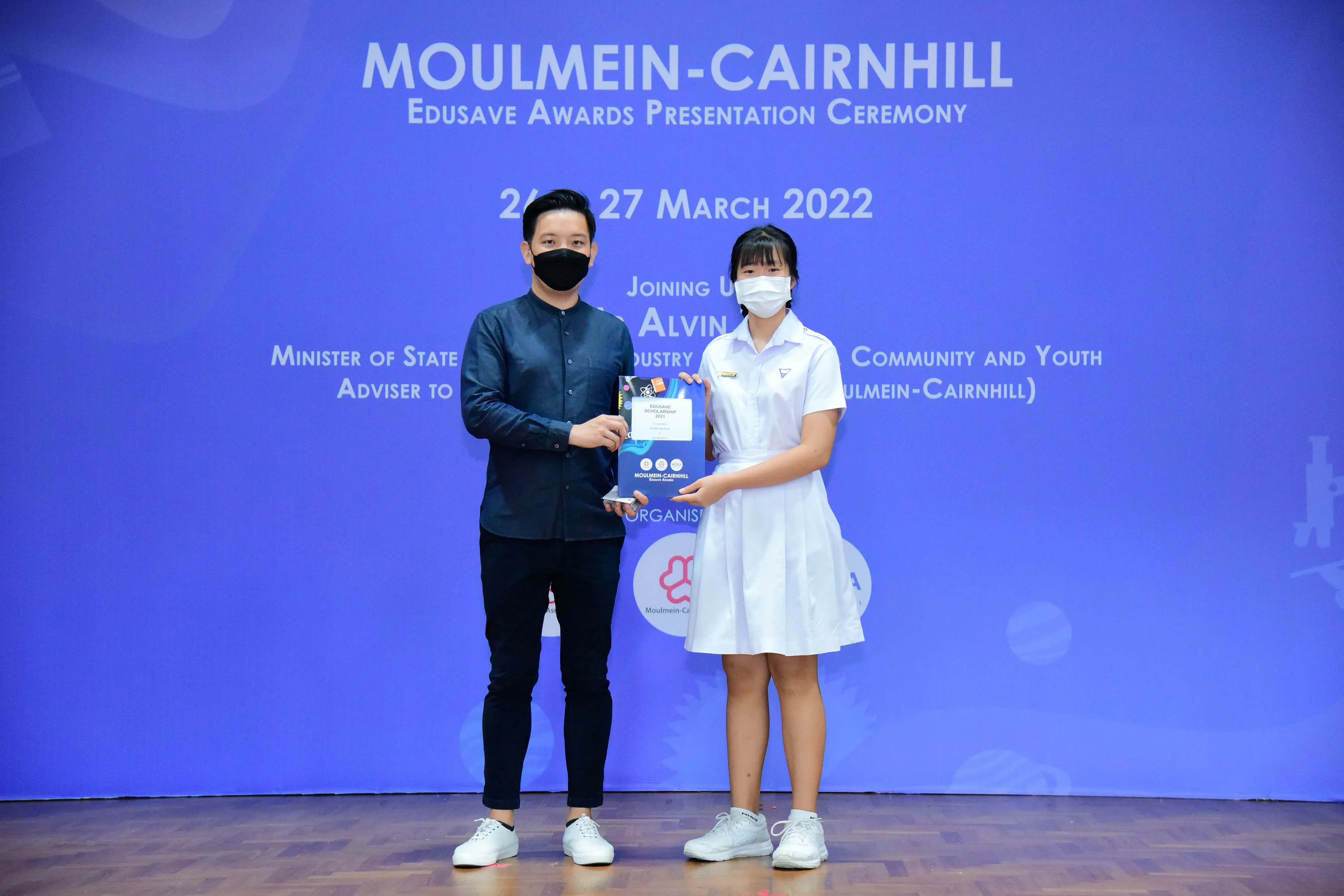 Two masked people hold an award at the Moulmein-Cairnhill Edusave Awards Presentation Ceremony, March 2022.