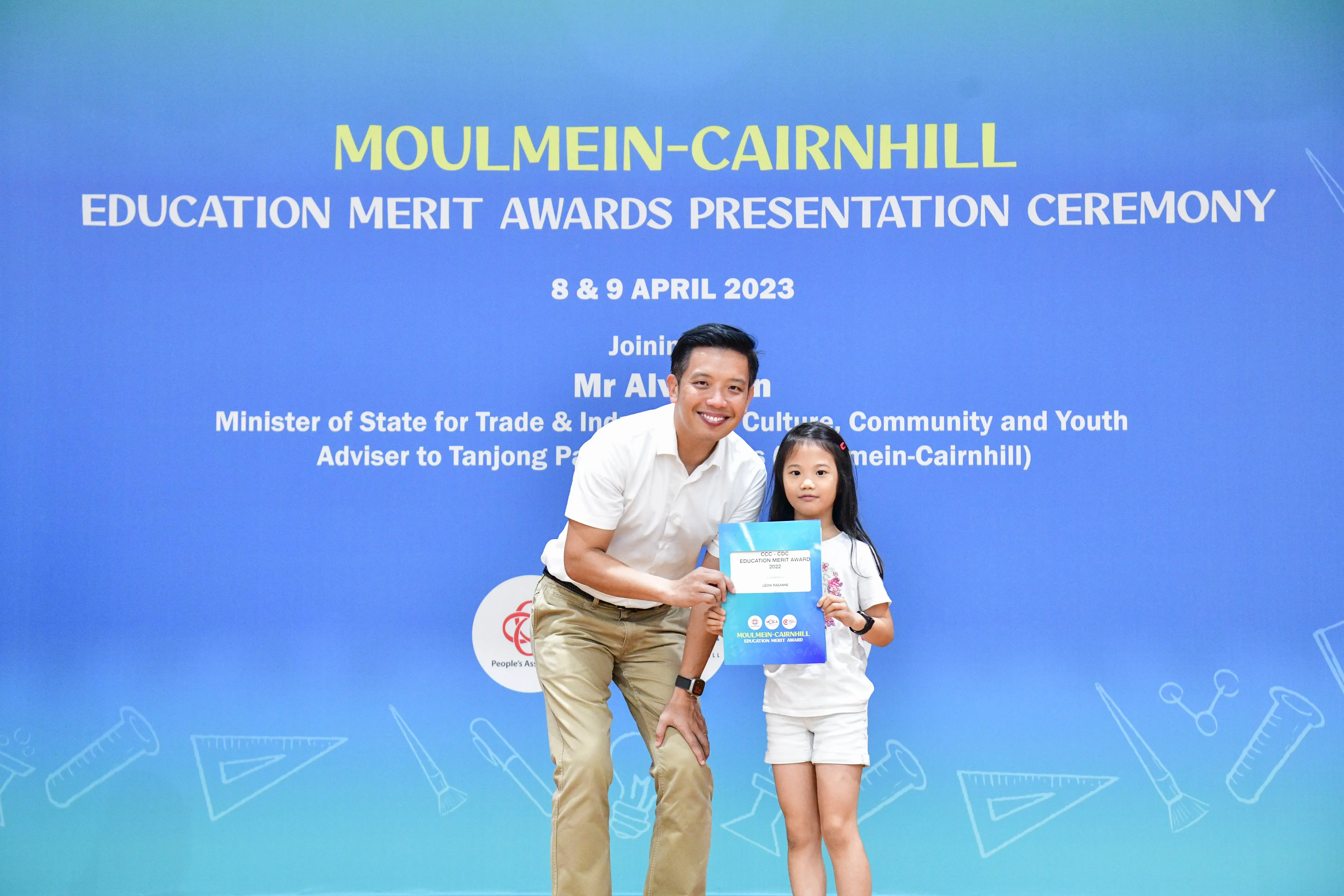 A man and a young girl hold a certificate in front of a Moulmein-Cairnhill Education Merit Awards banner dated April 2023.