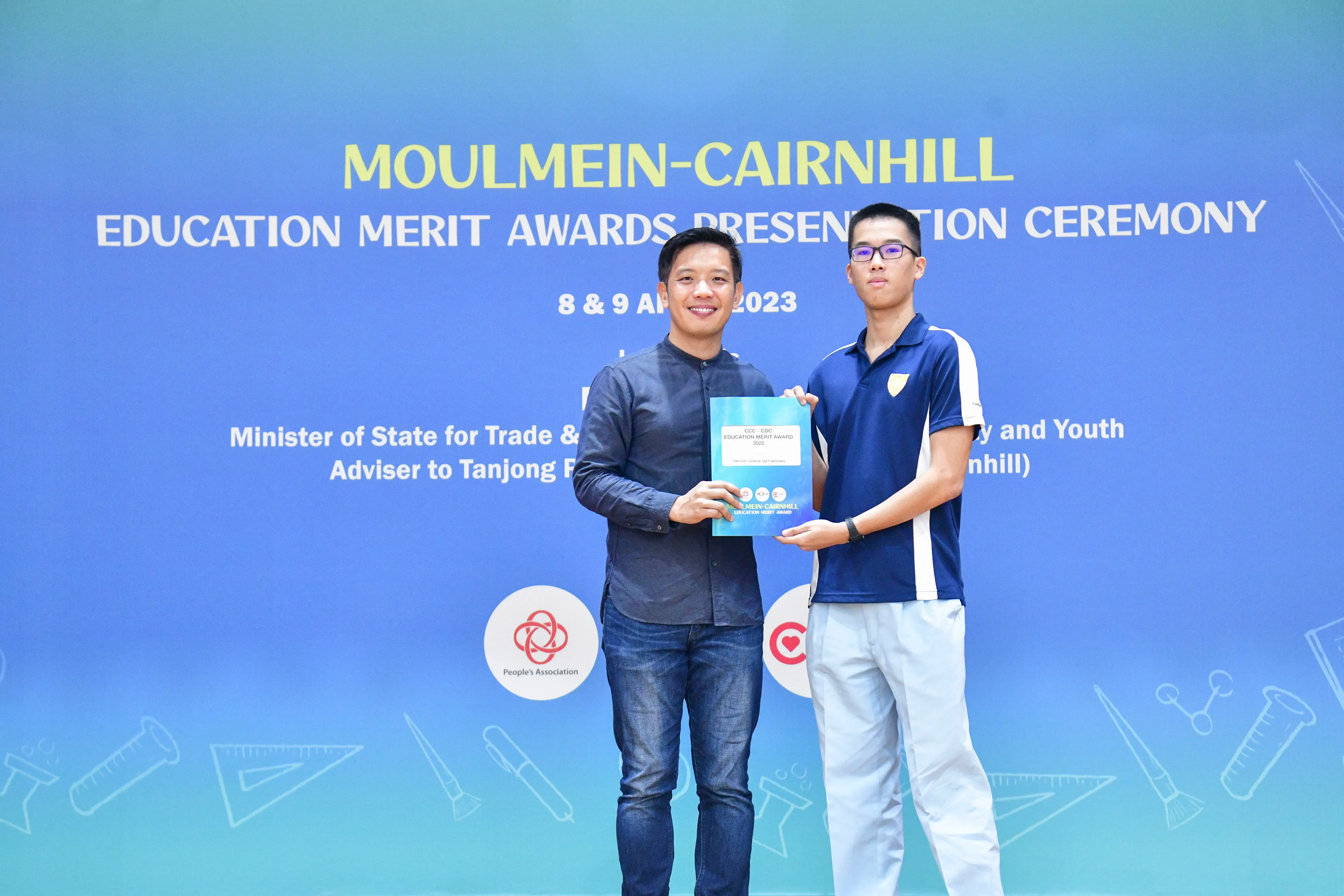 Two men hold a certificate together in front of a blue Moulmein-Cairnhill Education Merit Awards banner dated 2023.