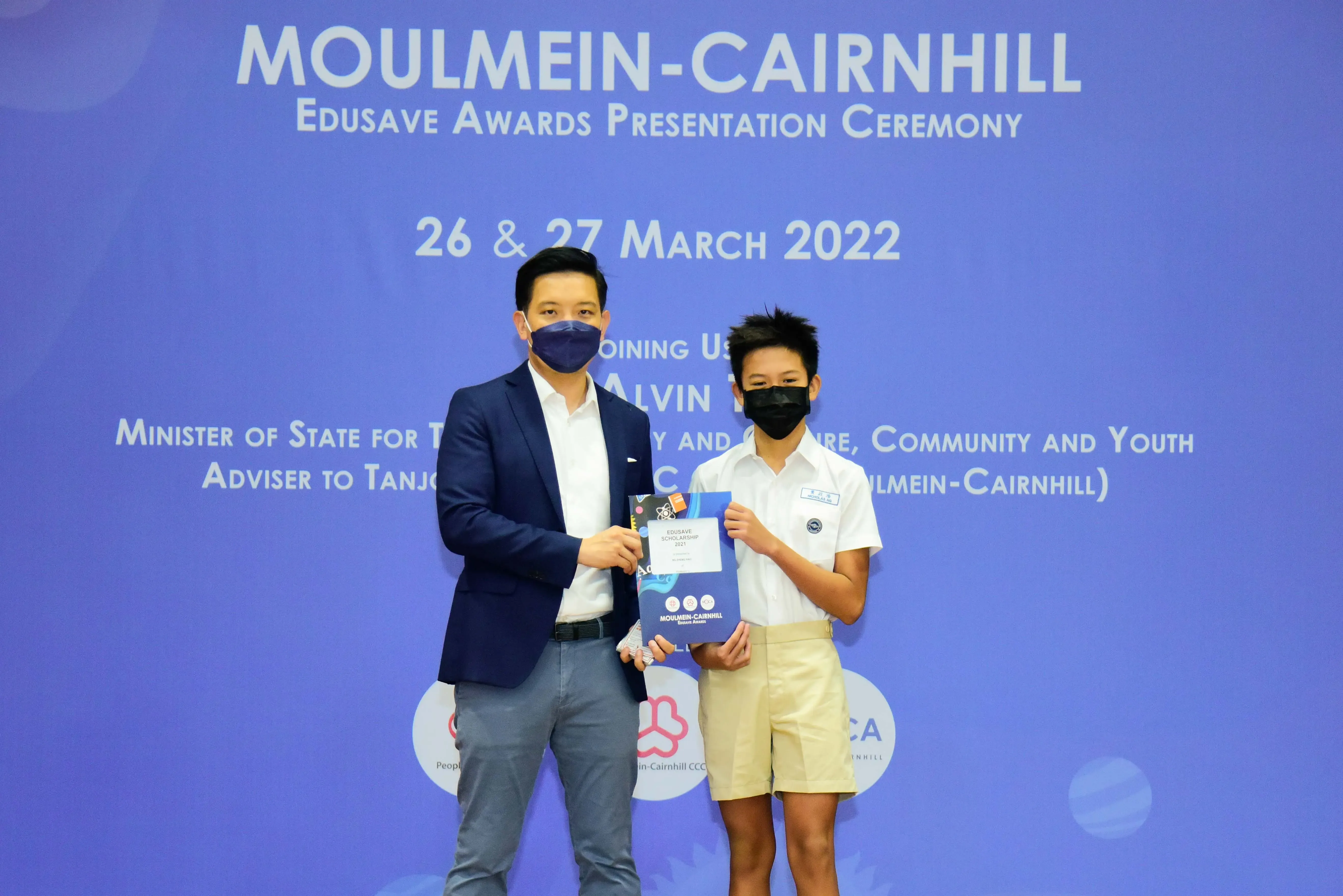 Adult and young student in masks hold an award at Moulmein-Cairnhill Edusave Awards Ceremony, March 2022.