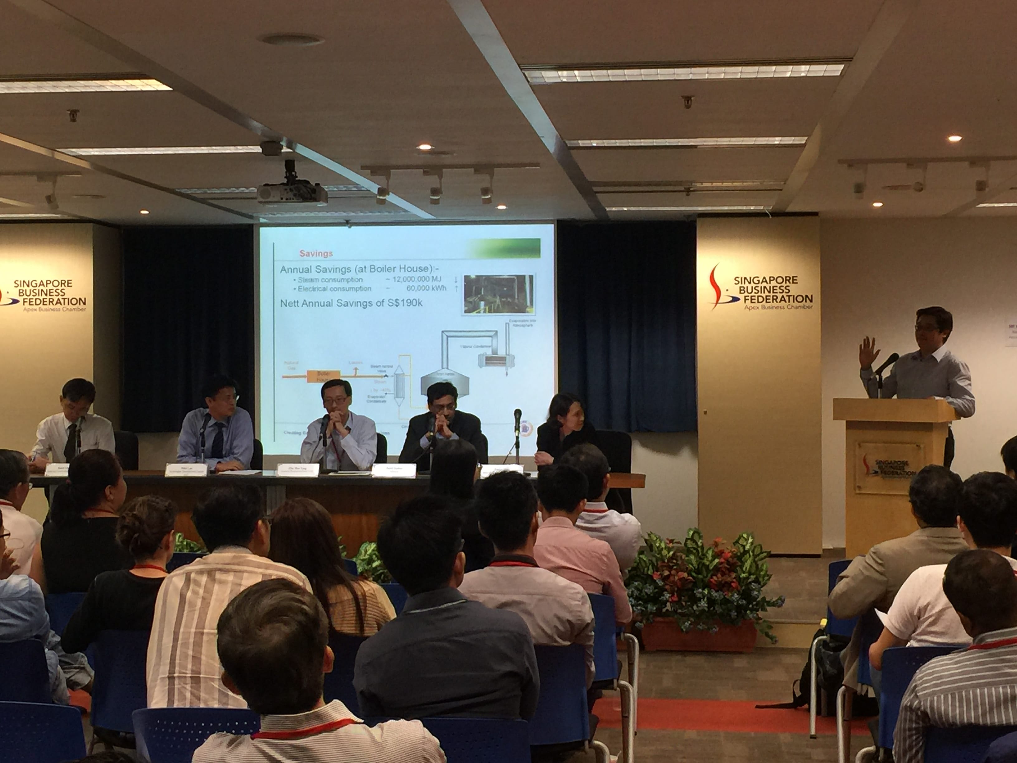 A speaker at a podium addresses an audience at a Singapore Business Federation event with a savings presentation on screen.