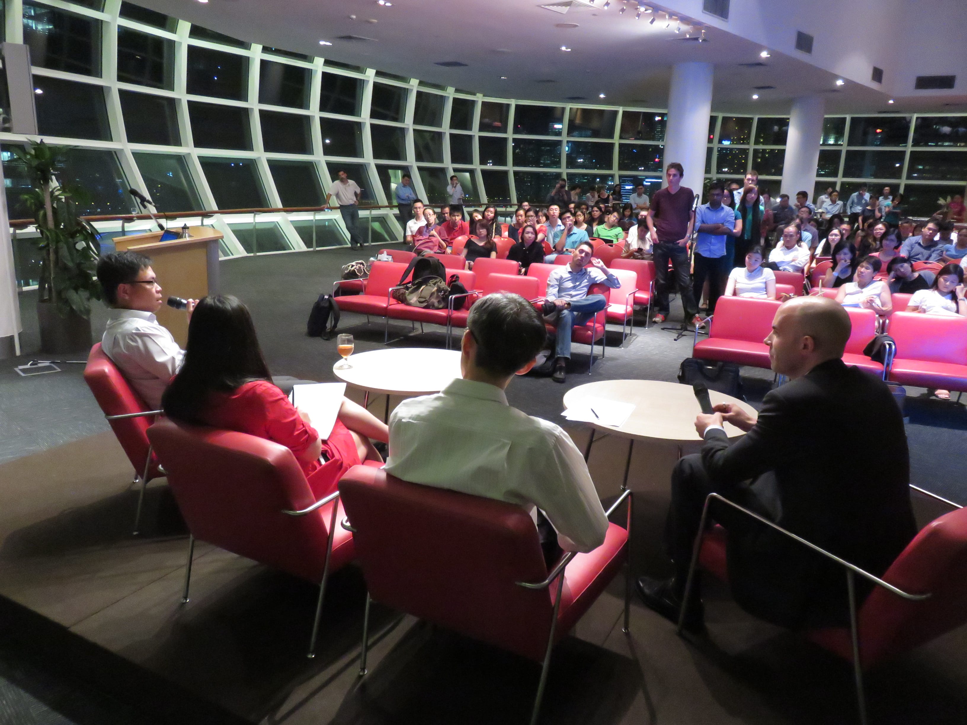 A panel of four people sits in red chairs facing a large audience in a modern venue with floor-to-ceiling windows at night.
