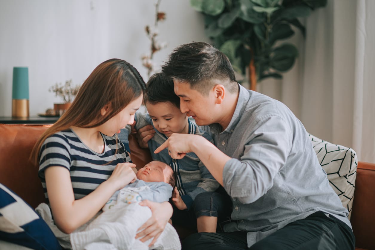 A woman holds a newborn on a sofa while a man and young boy lean in closely to look at the baby.