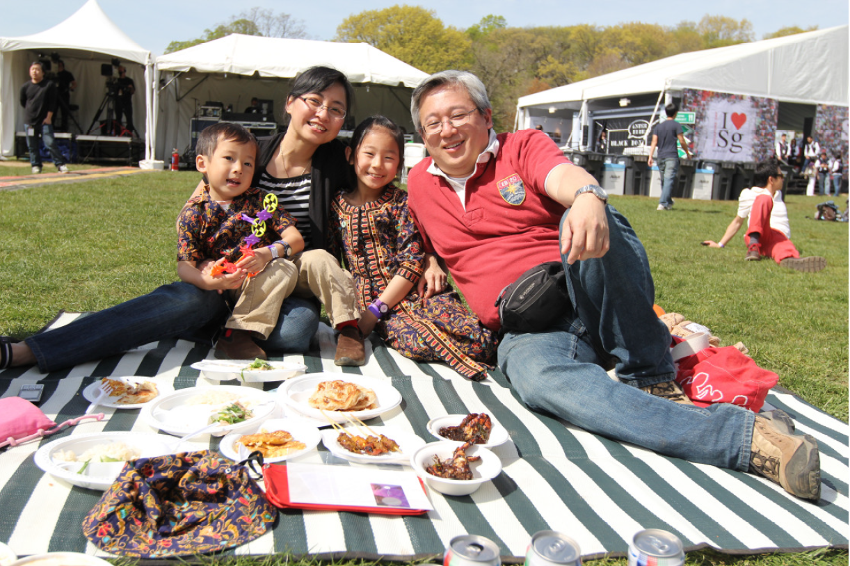Family of four sitting on a striped blanket outdoors with plates of food, smiling at camera near white event tents.