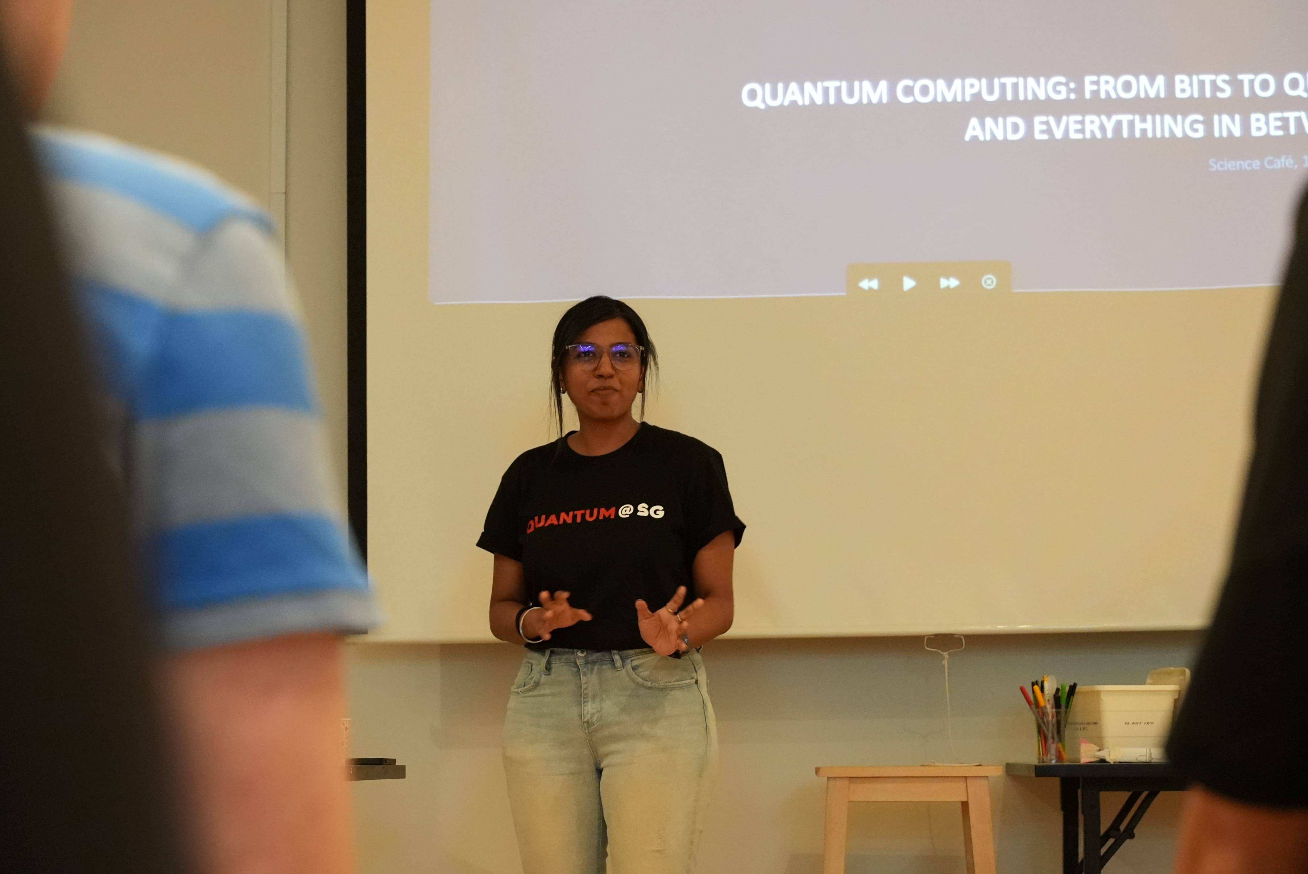 Woman in a black Quantum@SG shirt presenting in front of a projected quantum computing slide.