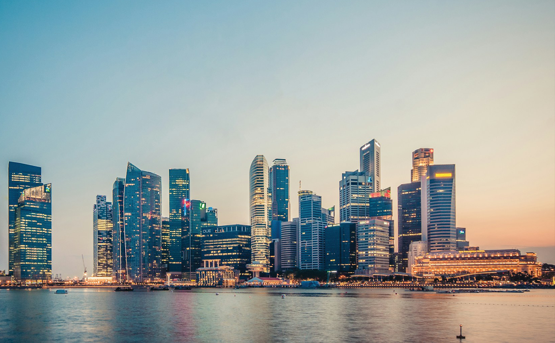 Singapore's glowing skyline with tall glass skyscrapers reflects over calm water at dusk under a pastel sky.