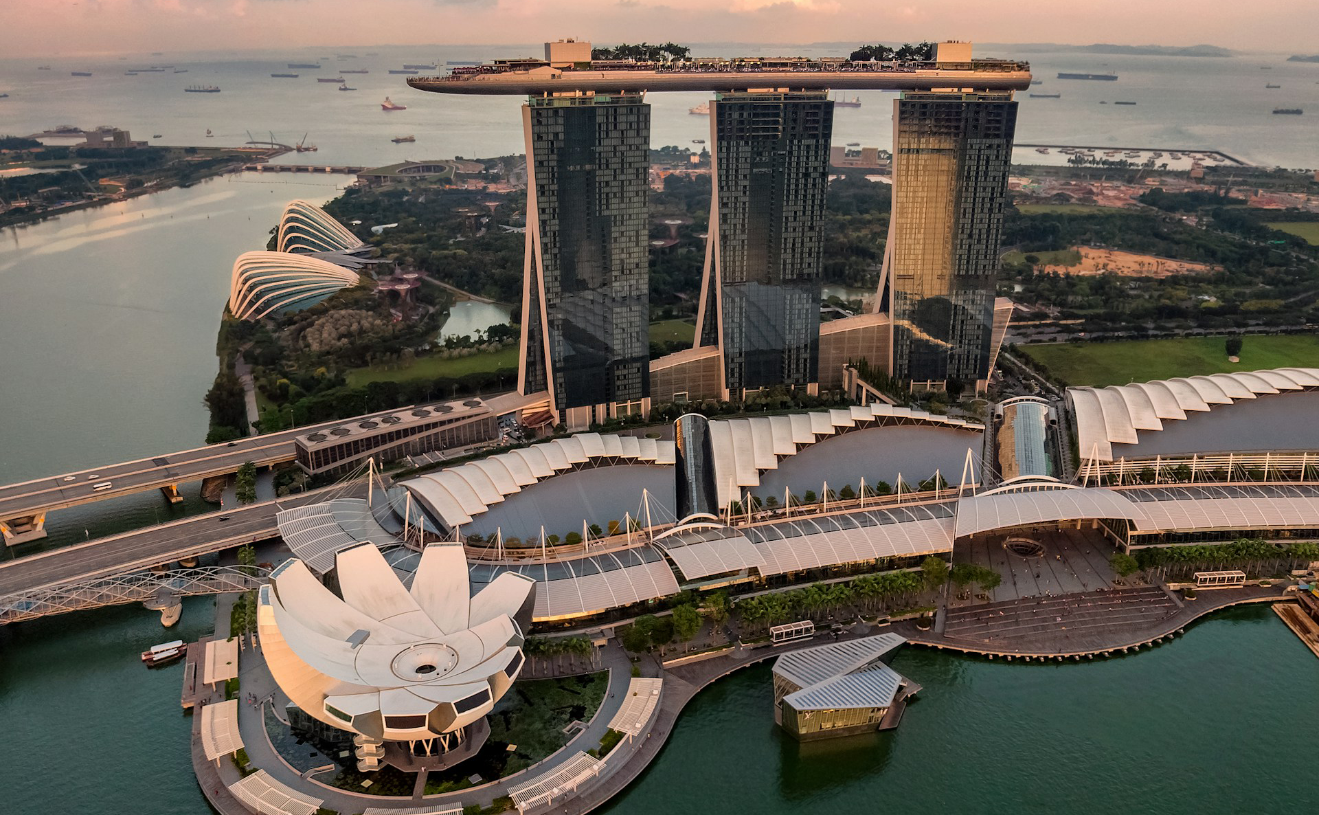 Aerial view of Marina Bay Sands hotel with three towers and rooftop deck, flanked by a lotus-shaped museum at sunset.