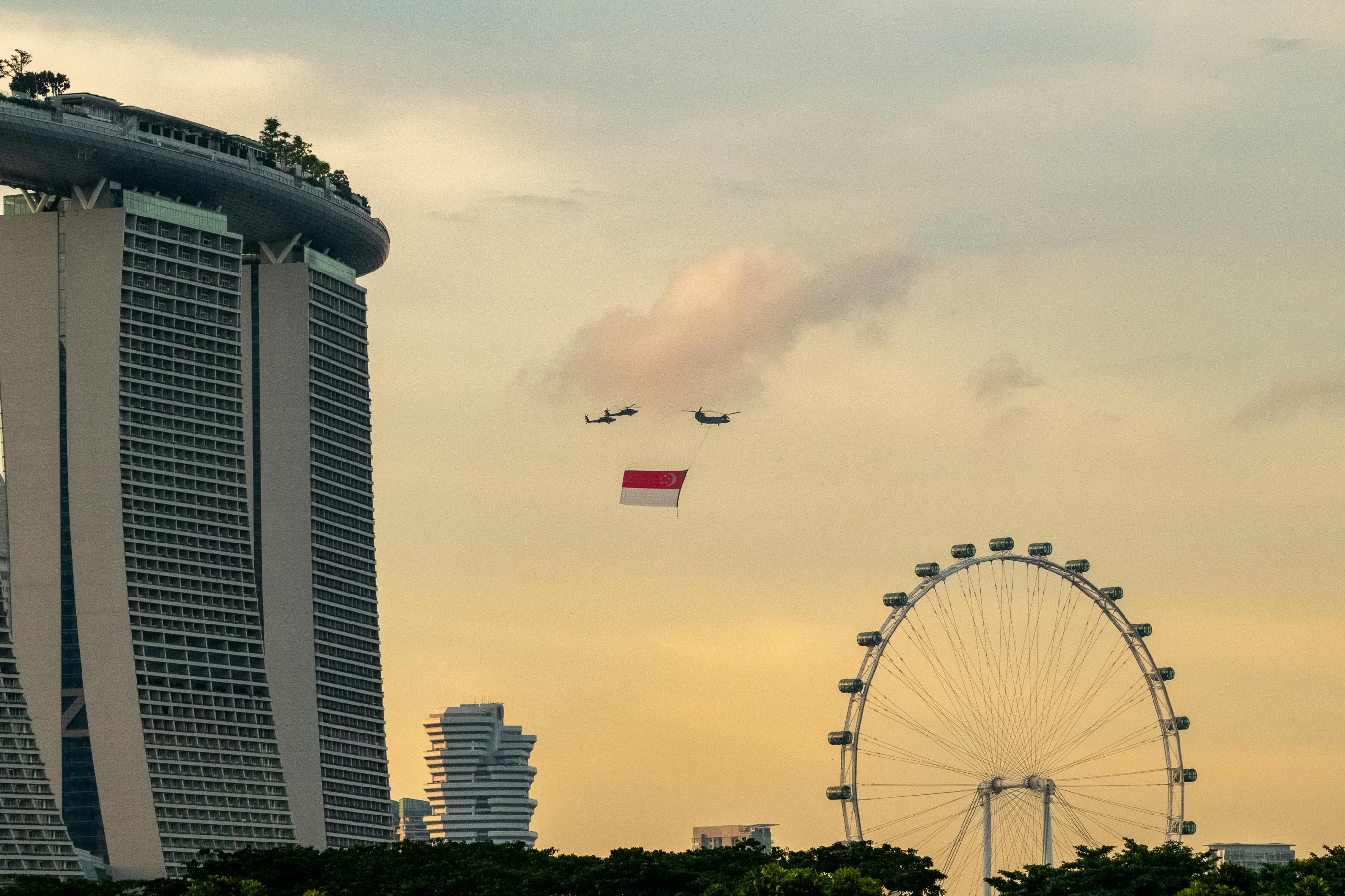 Helicopters carry a red and white flag over Singapore's skyline, with Marina Bay Sands and a Ferris wheel visible.