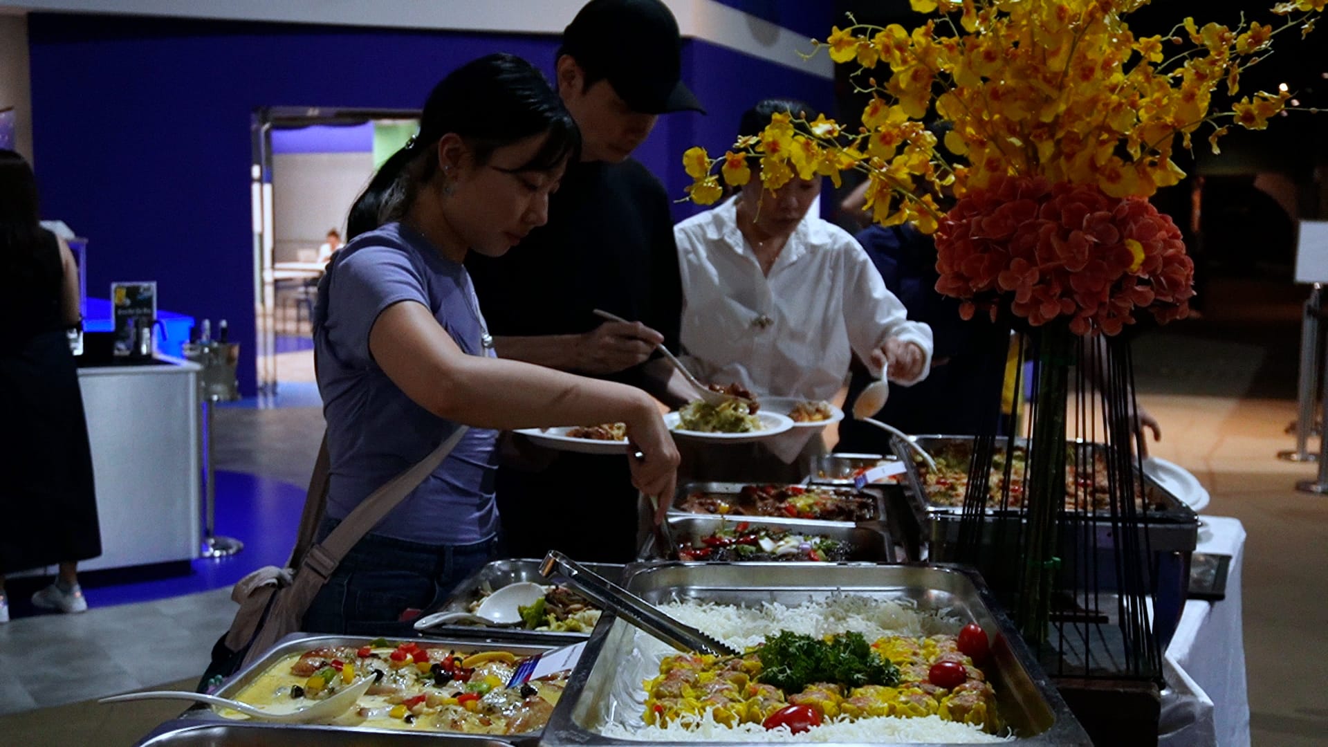 People serving themselves at a buffet with multiple food trays and a large yellow and red flower arrangement nearby.