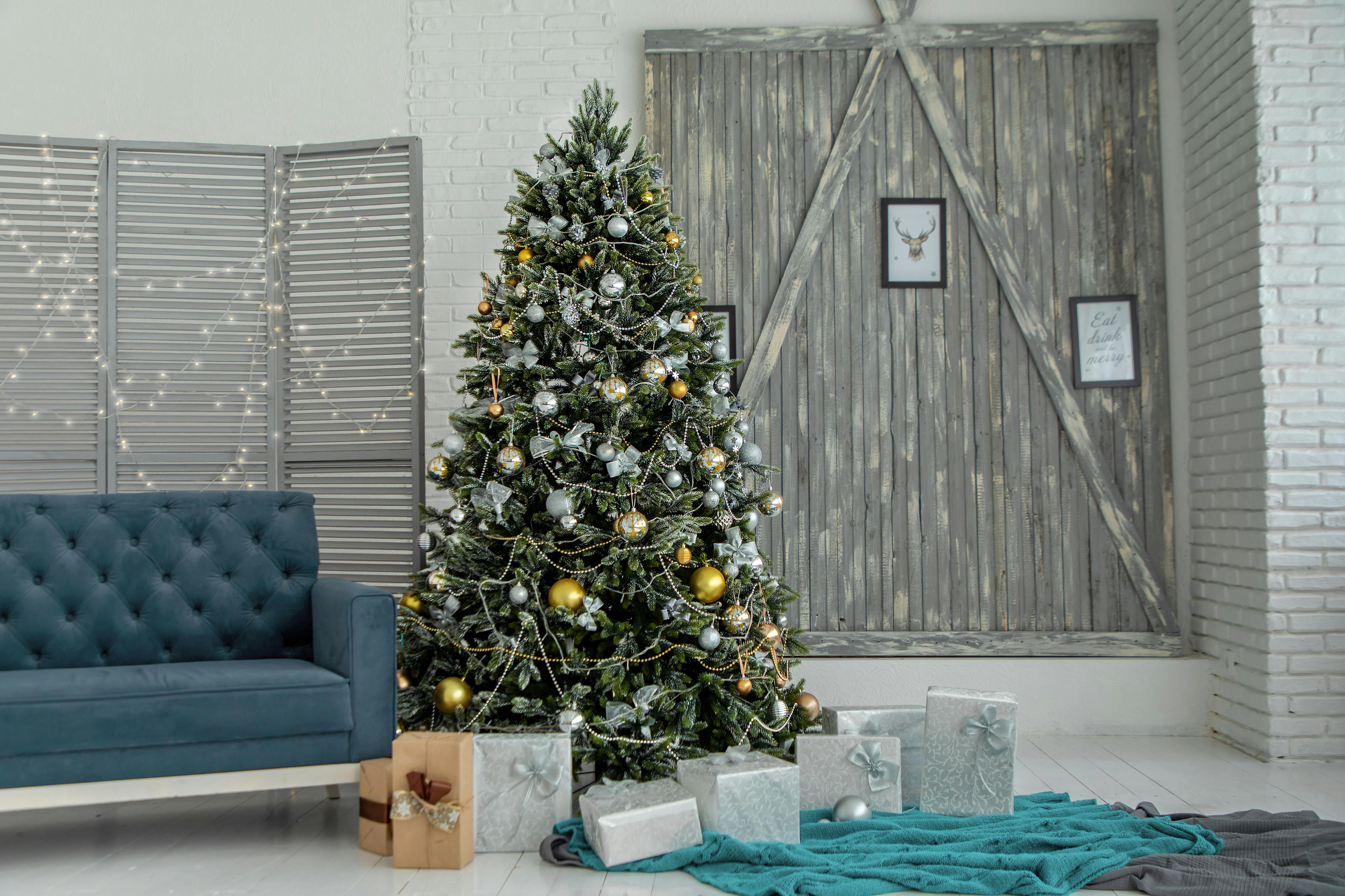 Decorated Christmas tree with gold and silver ornaments, surrounded by wrapped gifts, beside a blue sofa in a bright room.