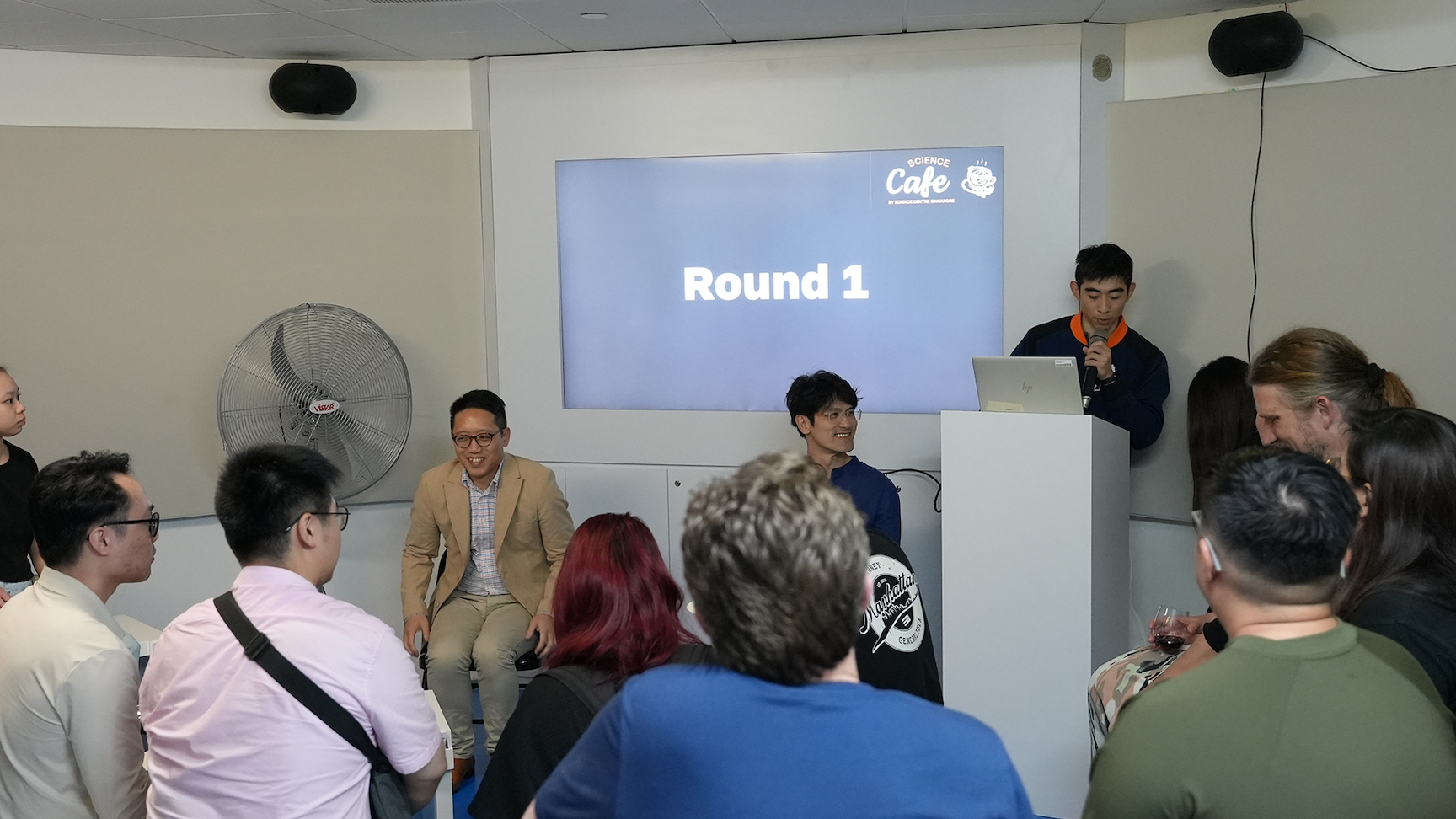 A host speaks at a podium with a screen showing "Round 1" at a Science Cafe event, with seated panelists and an audience.