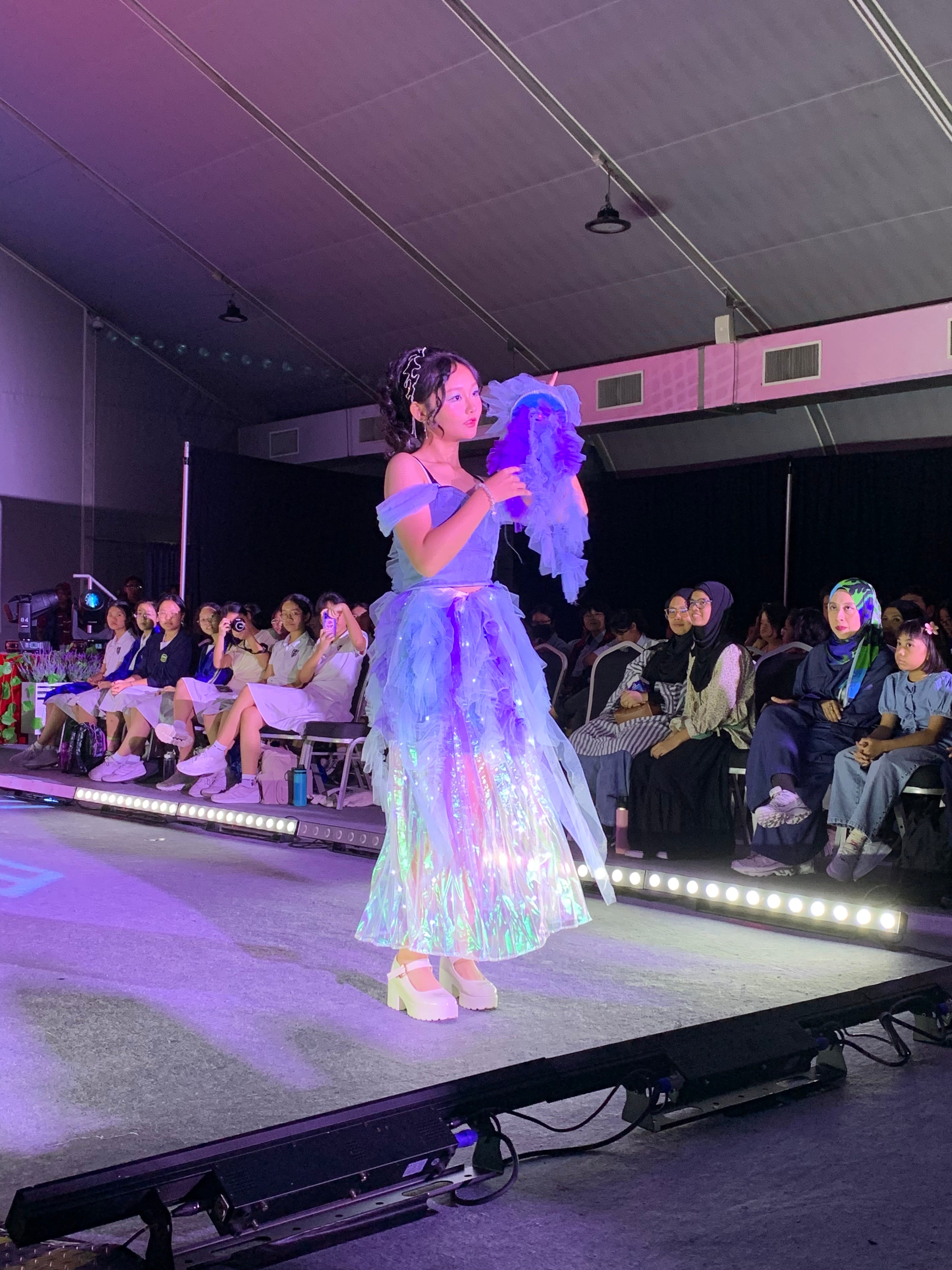 A young woman walks a runway in a purple iridescent ruffled gown holding a feathery accessory, with seated audience on both sides.