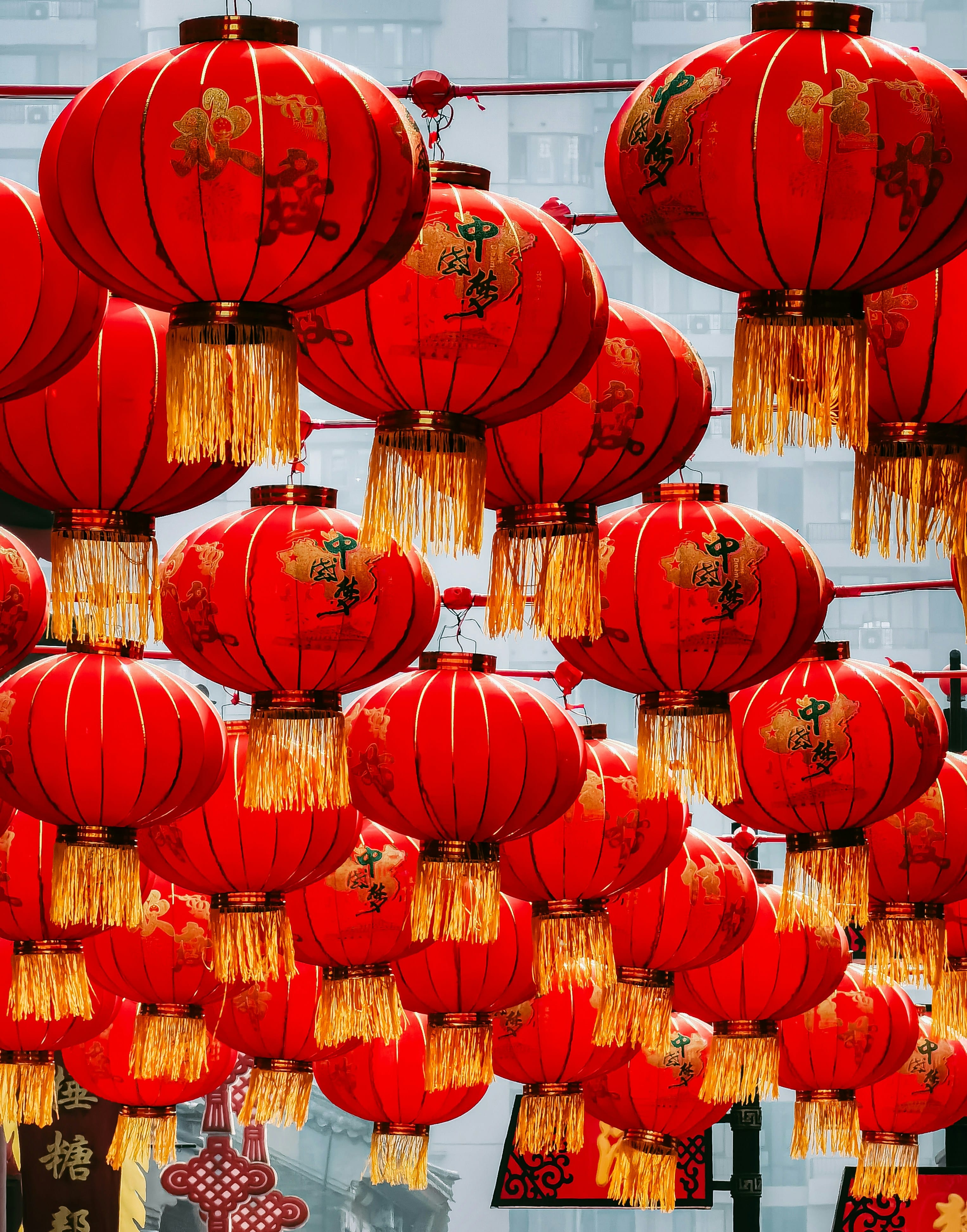 Rows of large red Chinese lanterns with gold tassels and Chinese characters hang overhead against a city backdrop.