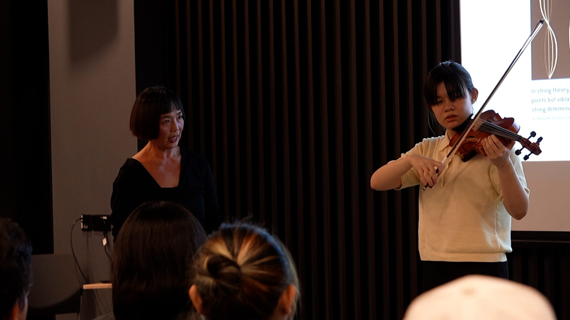 A young girl plays violin while a woman stands beside her, performing before a seated audience near a presentation screen.