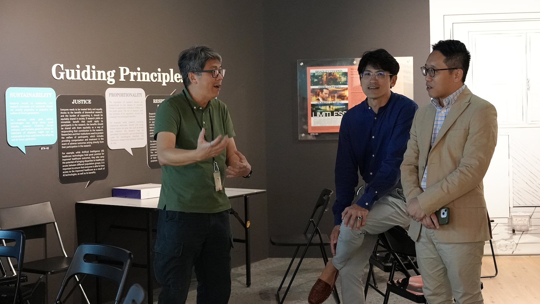 Three men converse in a room with a dark wall displaying a "Guiding Principles" exhibit with text panels.
