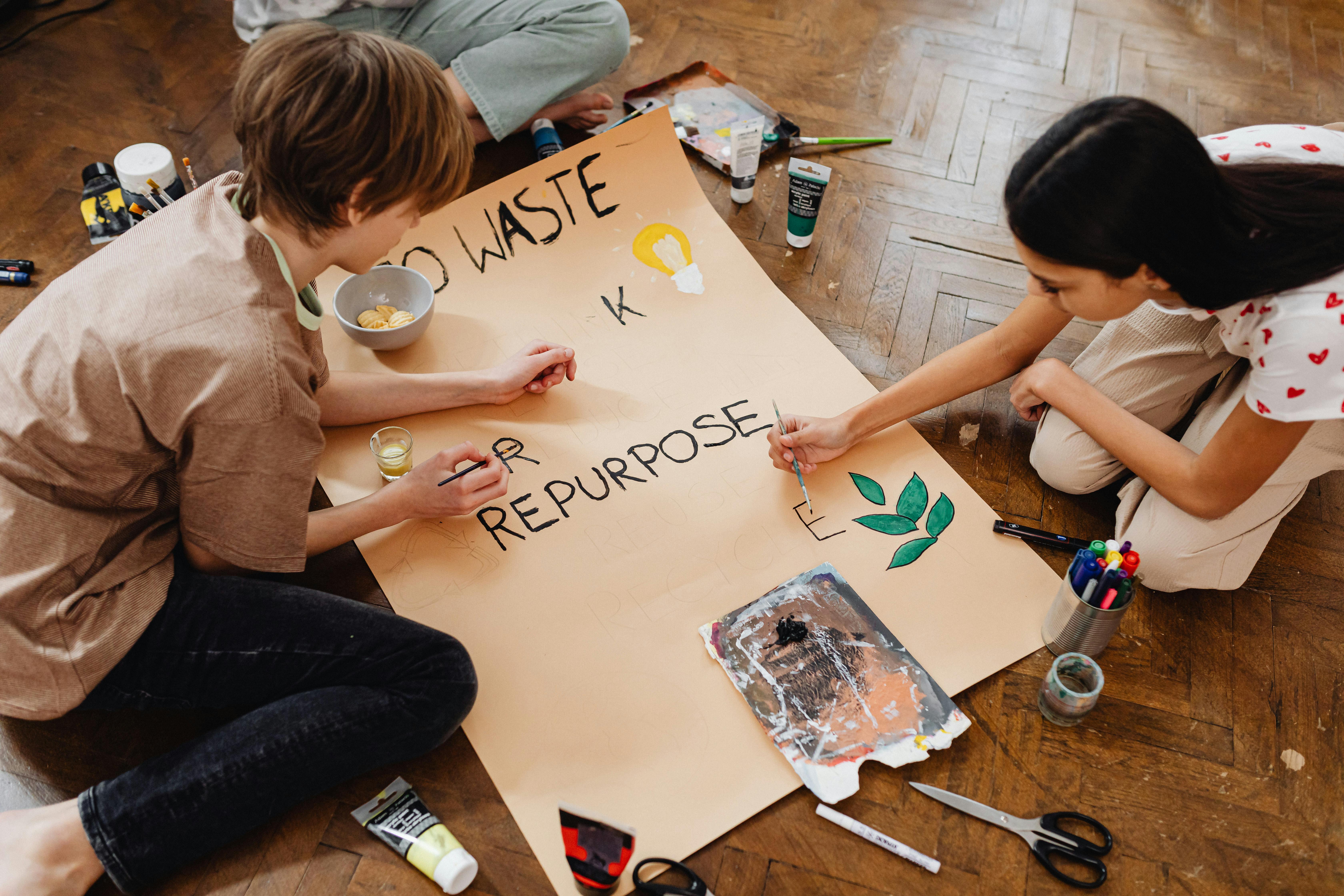 Two kids sit on a wood floor painting a large poster with "No Waste, Repurpose" and leaf and bulb illustrations.