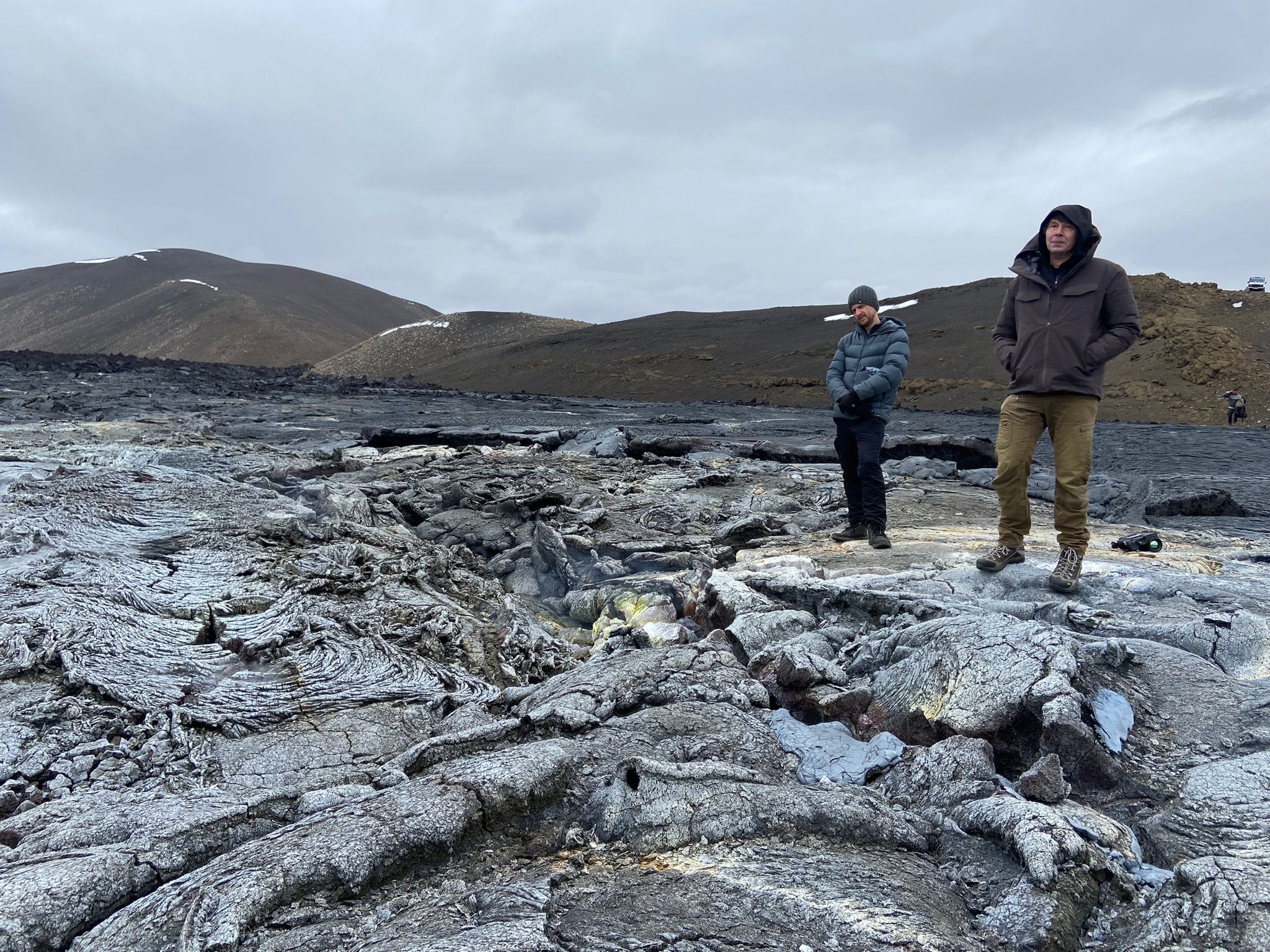 Two people in winter jackets stand on a vast dark hardened lava field with volcanic hills under a cloudy sky.