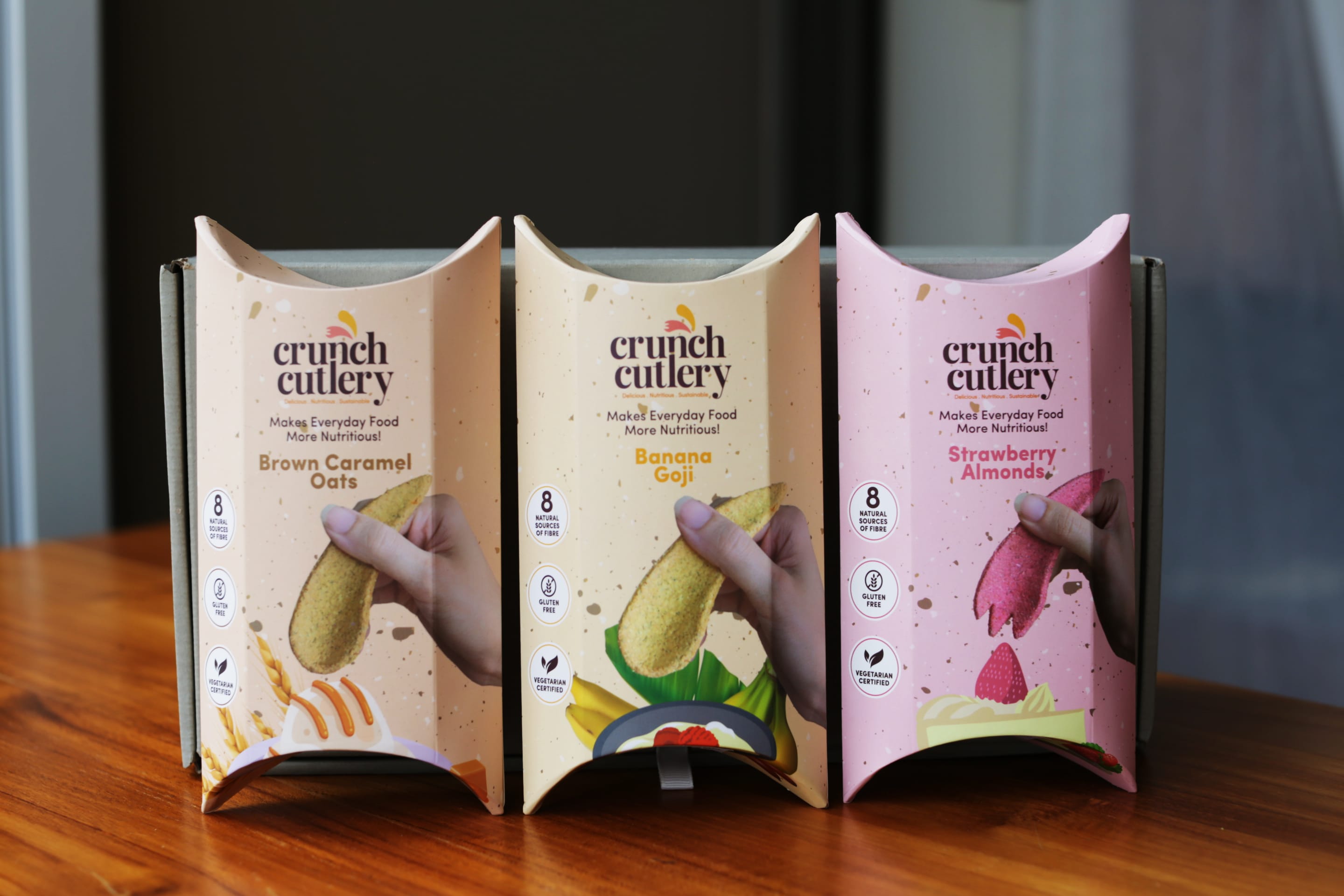 Three Crunch Cutlery pillow boxes in Brown Caramel Oats, Banana Goji, and Strawberry Almonds flavors on a wooden table.