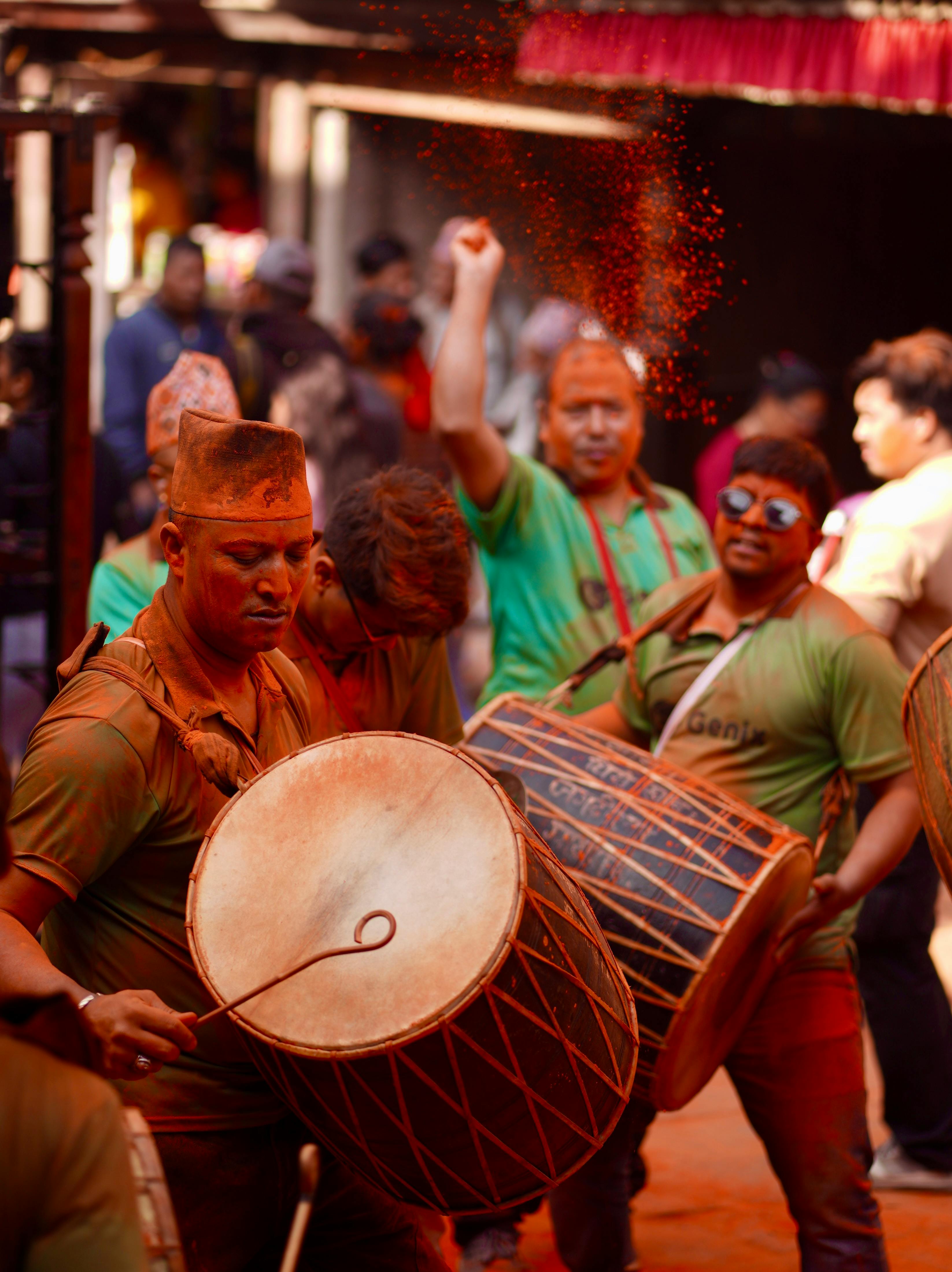 Men covered in red powder play traditional drums at a festive outdoor celebration with a crowd behind them.