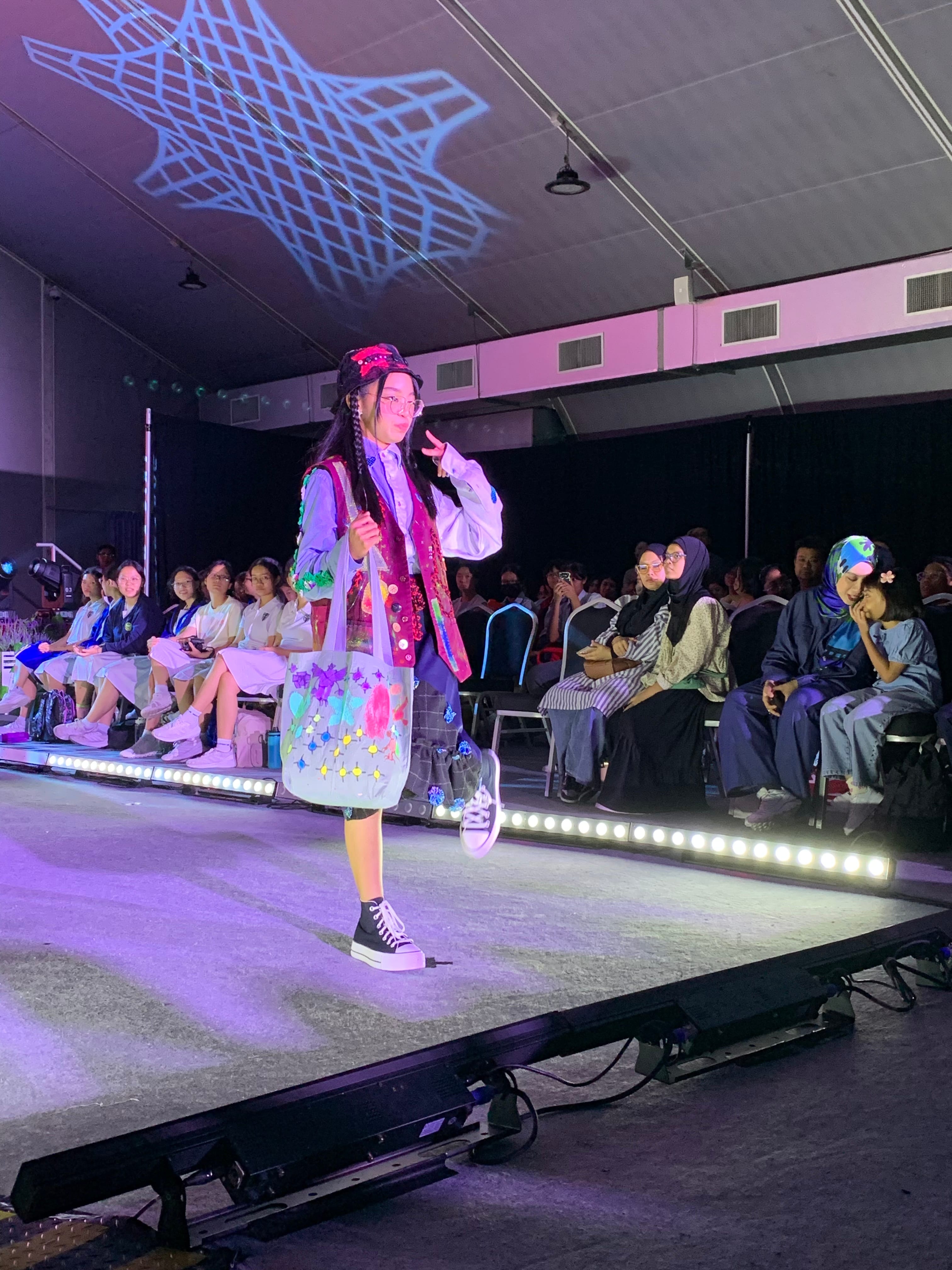 A young model walks a lit runway in colorful eclectic outfit with braids and decorated tote bag, audience seated on both sides.